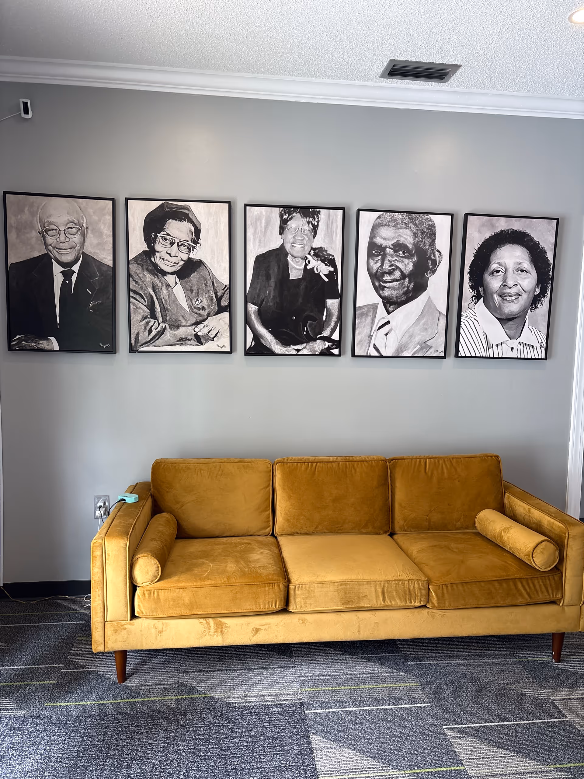 Lounge area with mustard sofa and five framed black-and-white portraits on a gray wall