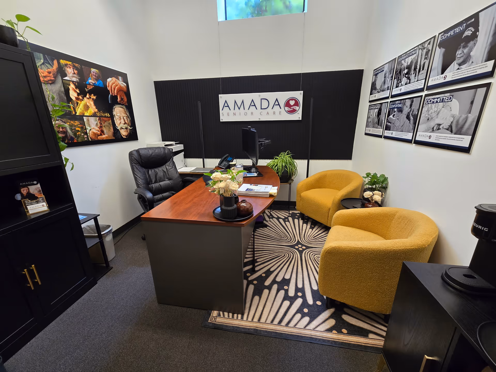 Office reception area featuring a desk, two yellow chairs, plants, and an Amada Senior Care sign.