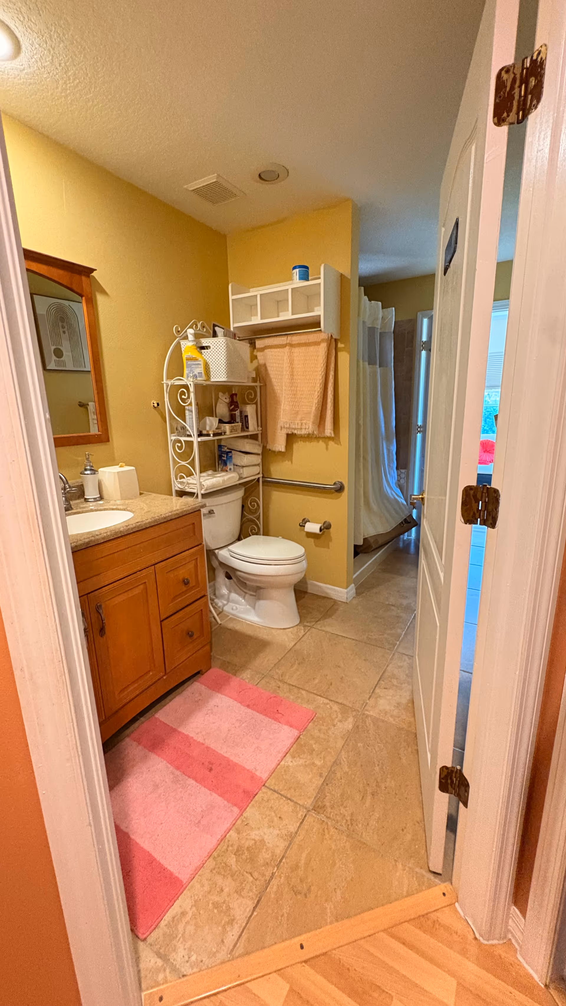 Yellow-walled bathroom with wooden vanity, toilet, towels, and a pink striped rug.