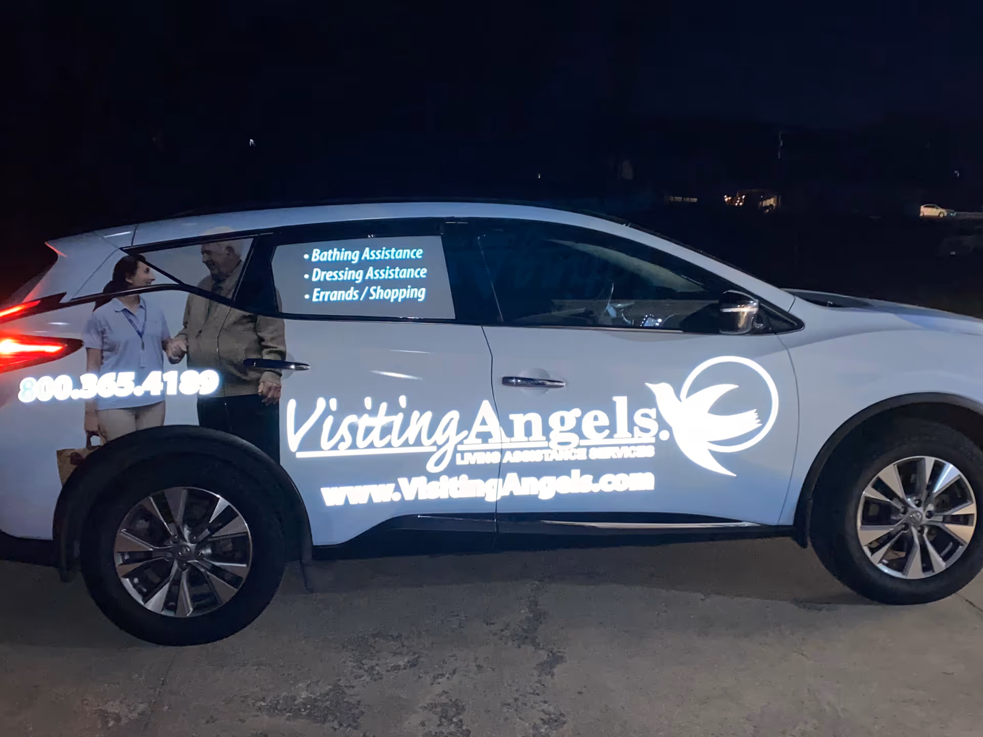 Nighttime side view of a Visiting Angels branded car with logo and two-person graphic