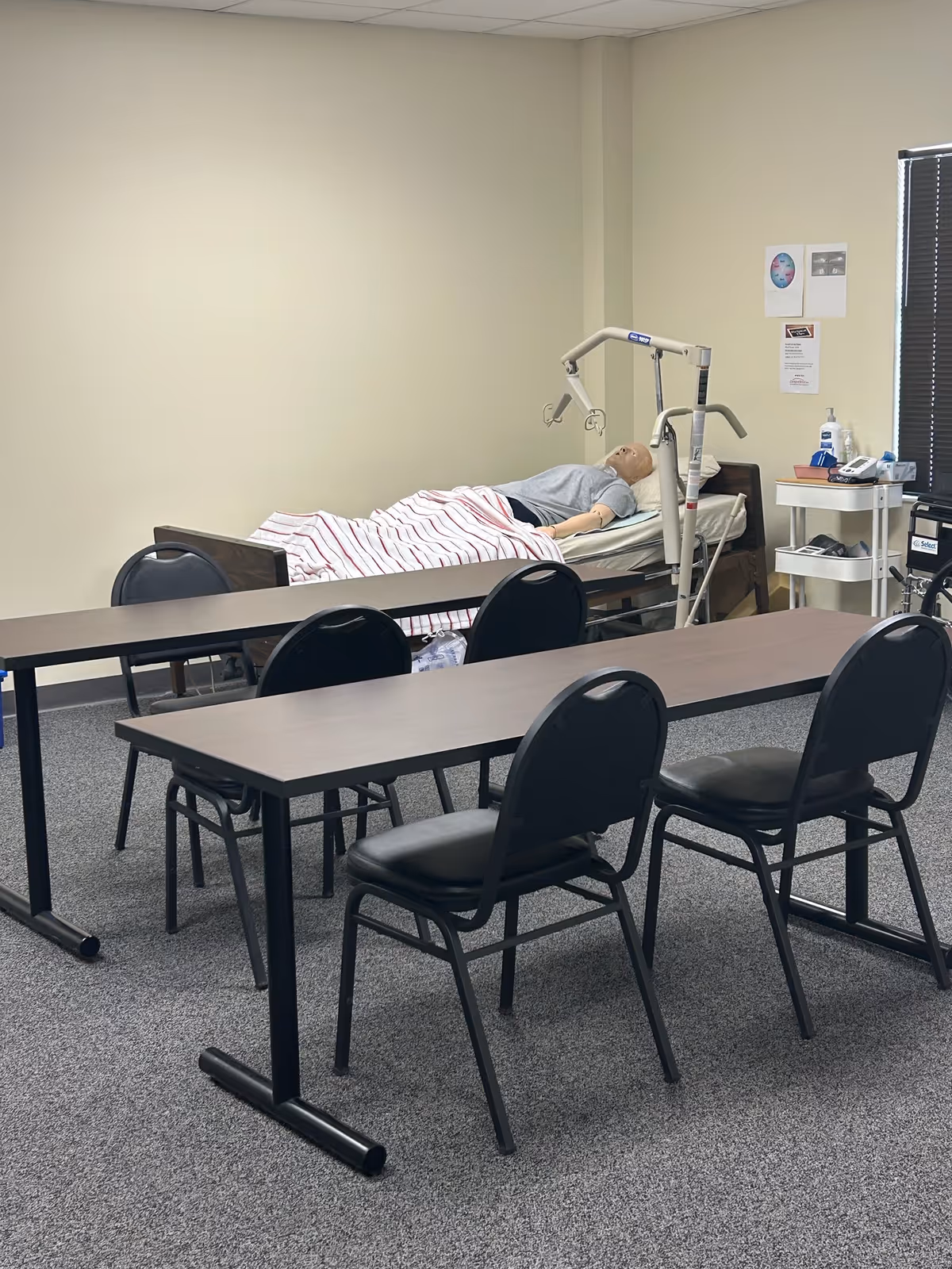 A resident lies on a hospital bed with medical equipment in a classroom-style room.