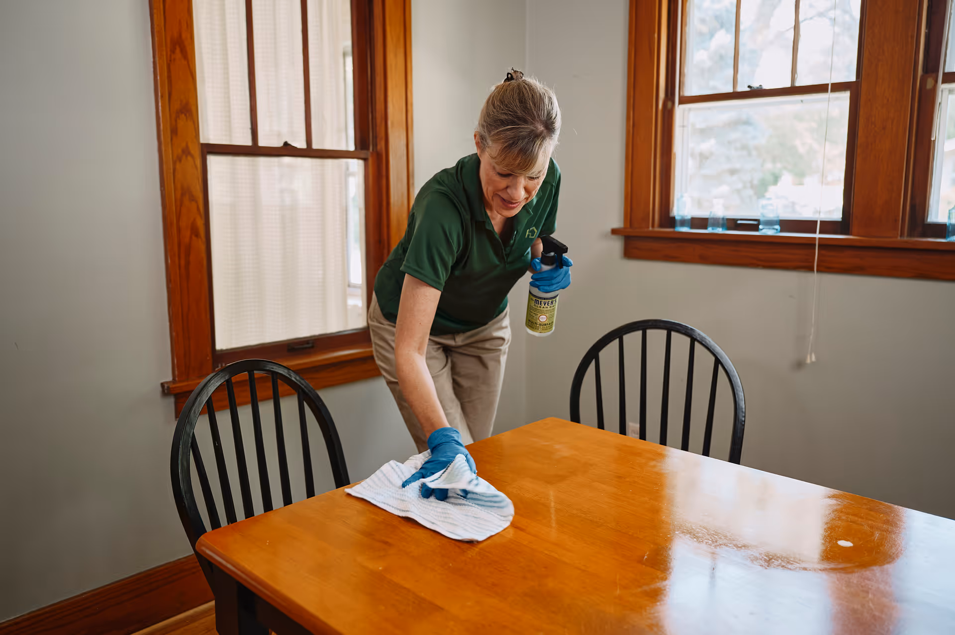 Caregiver cleans a wooden dining table in a bright dining room.