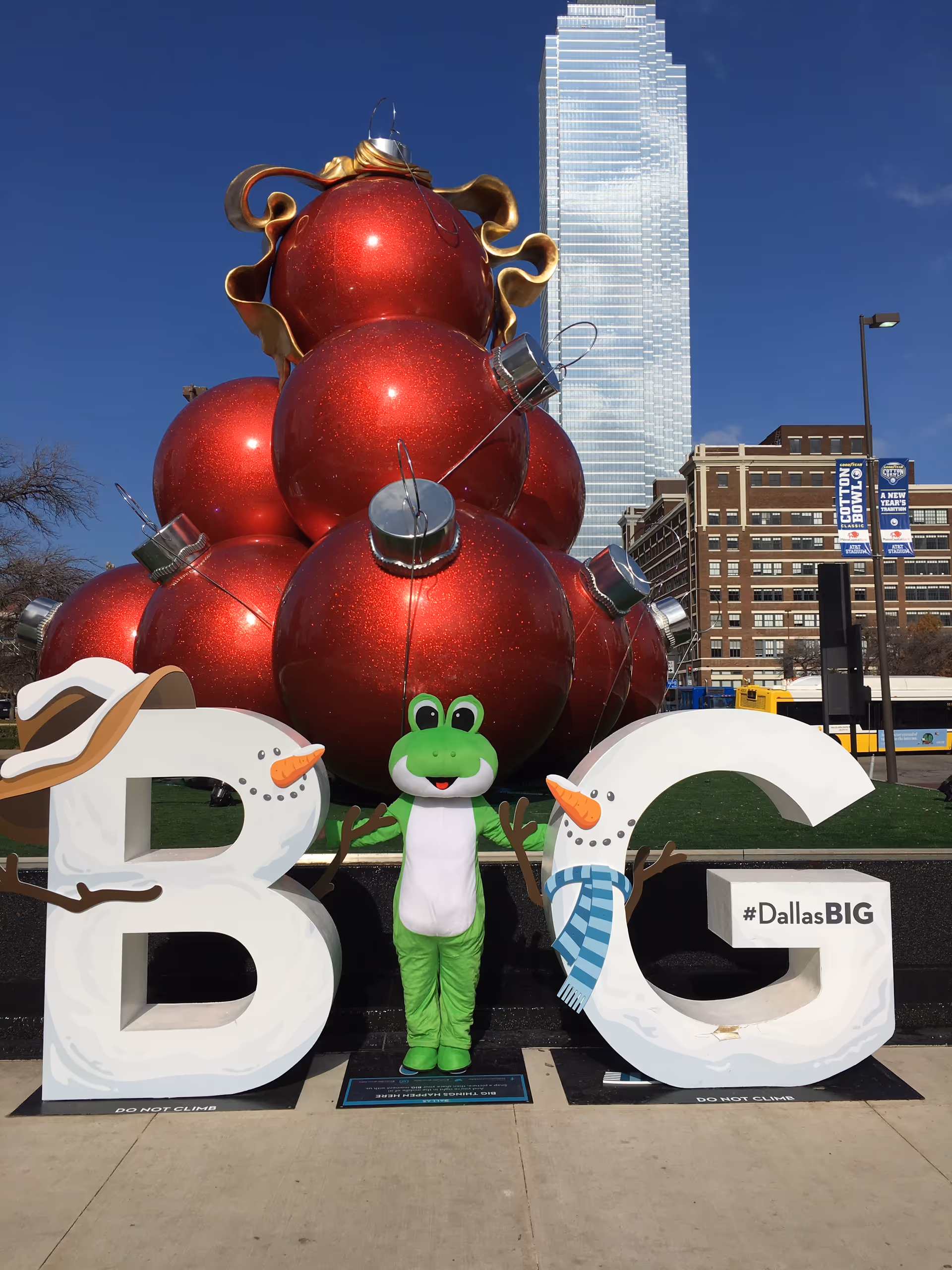 Costumed frog mascot stands between oversized B and G letters beside red ornament sculpture.