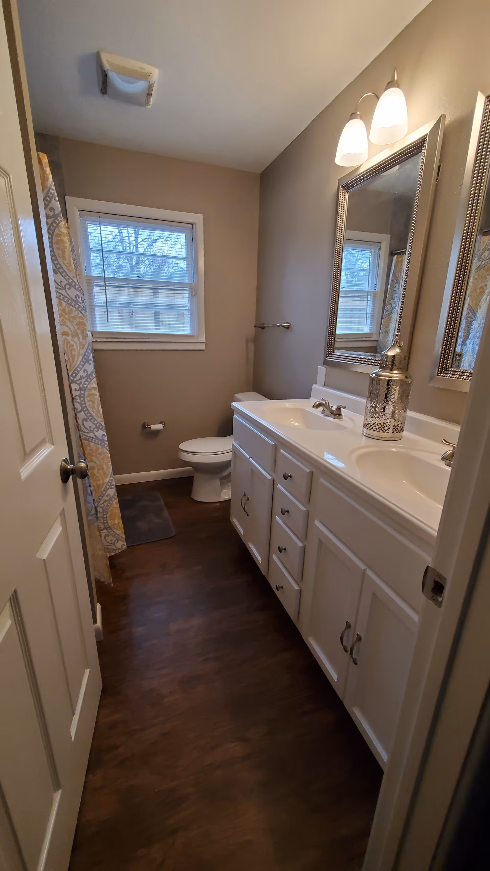 Compact bathroom with double sink vanity, toilet, window, shower curtain, and mirror.