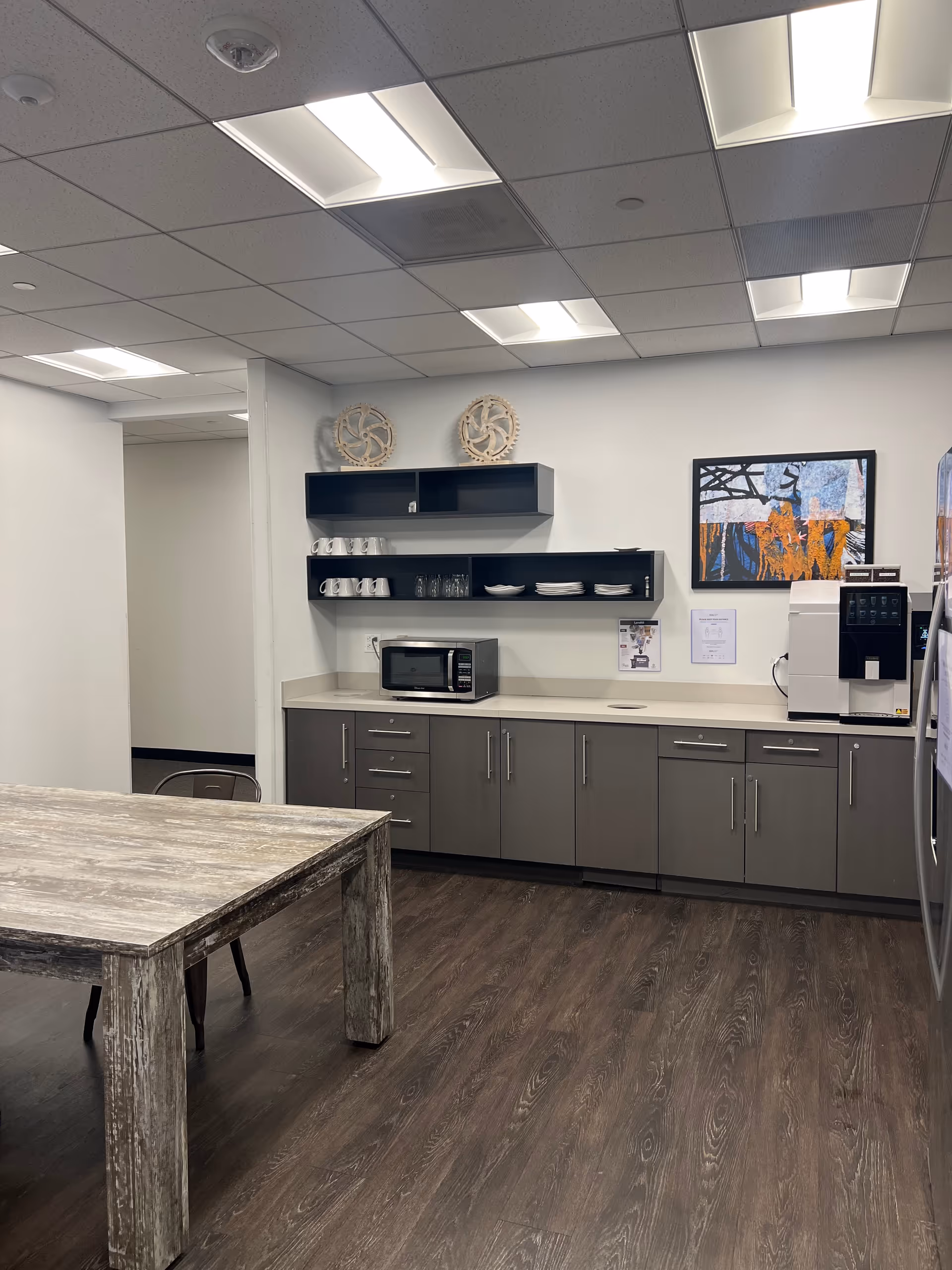 Modern staff kitchenette featuring microwave, coffee machine, open shelves, and a rustic table.