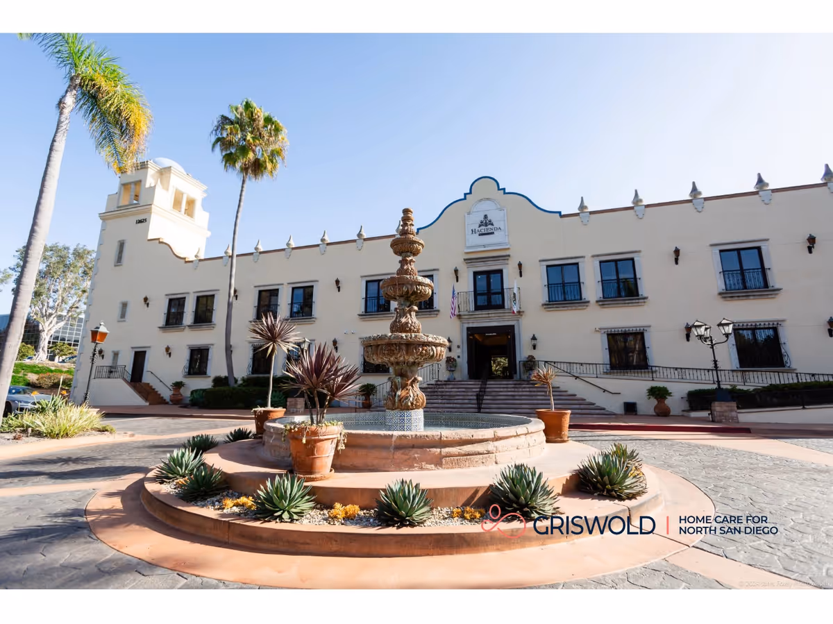 Exterior view of a light-colored building with a circular fountain in a palm-filled courtyard.