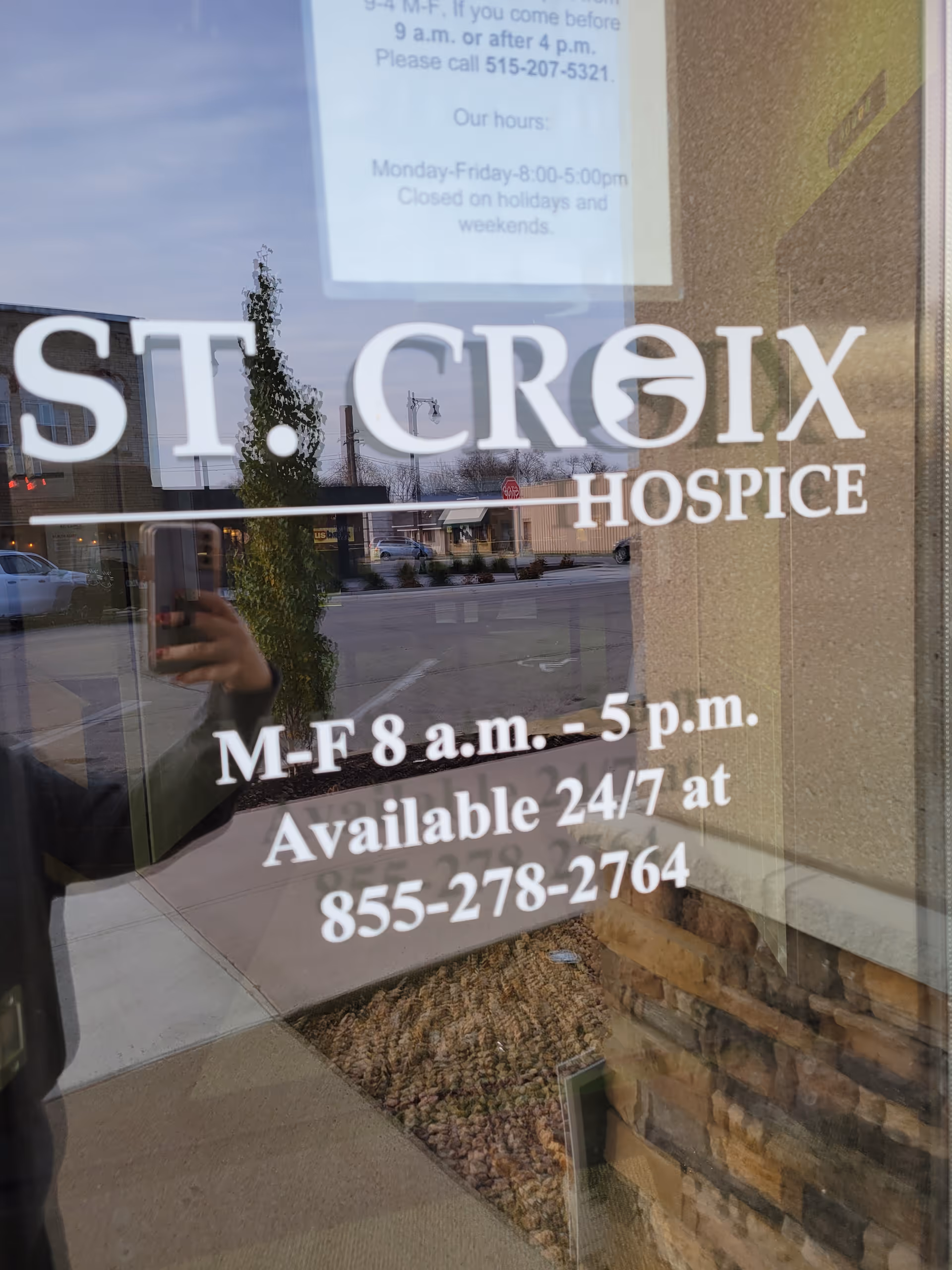 Glass door signage shows St. Croix Hospice hours; street is reflected outside.