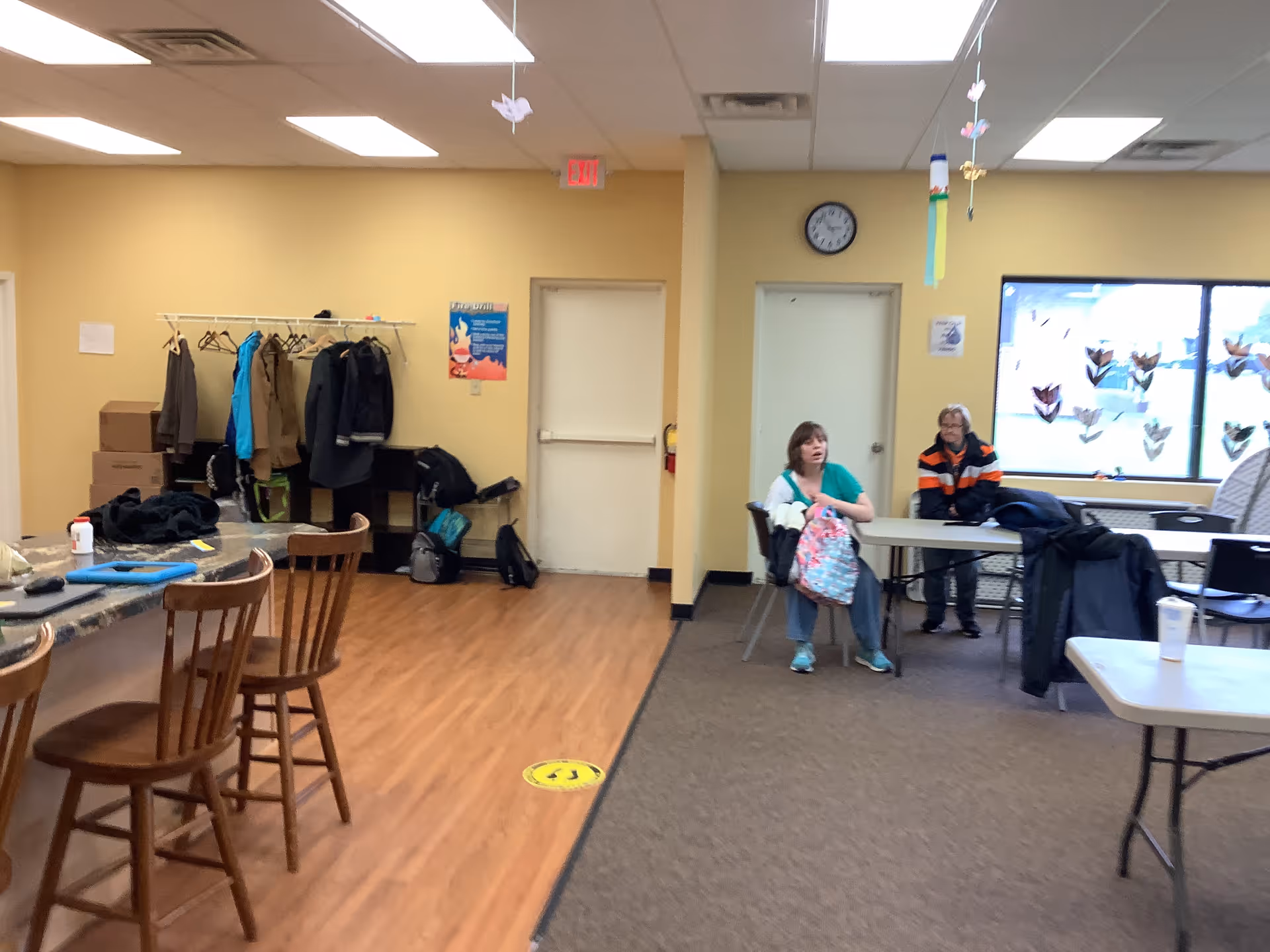 Interior common area in a senior facility with coat rack, tables, and two residents.