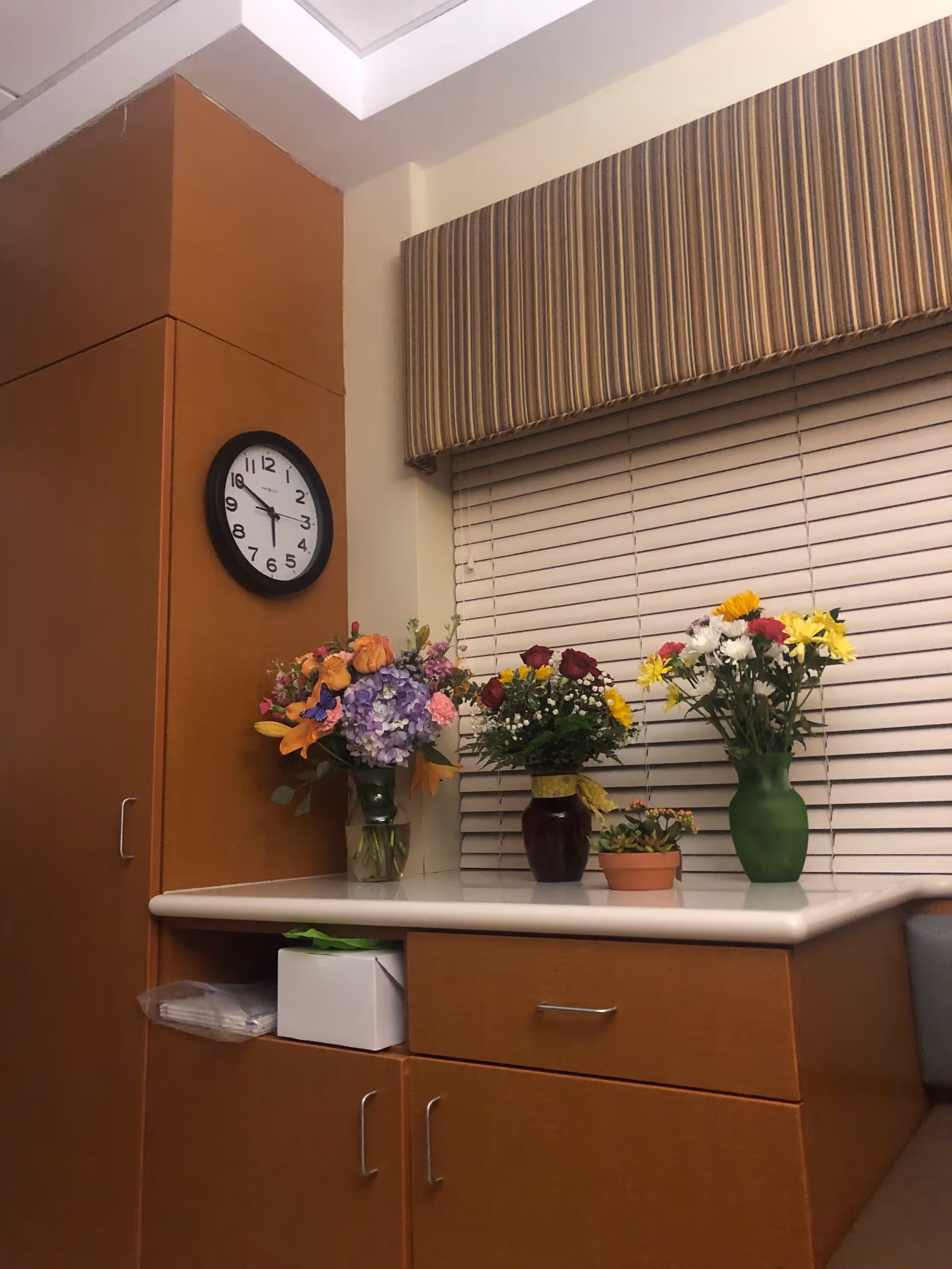 Interior corner with wooden cabinets, a clock, blinds, and vases of colorful flowers on a counter.