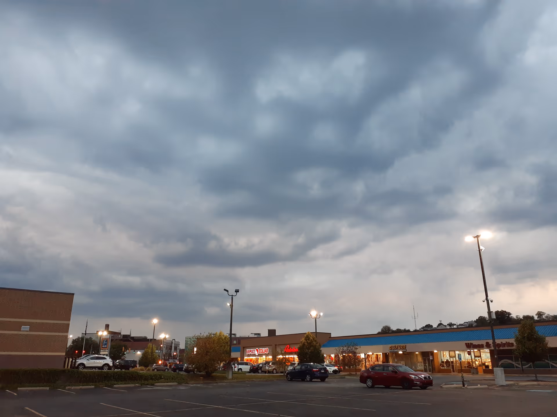 Overcast sky above a strip mall parking lot with storefronts and cars.