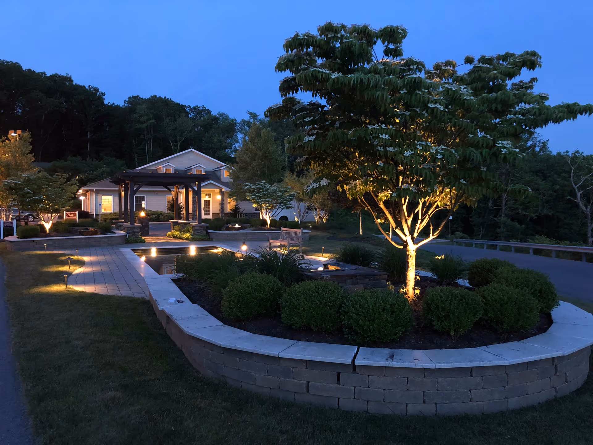 Evening exterior of a senior living facility with lit trees, circular garden, and a pathway.