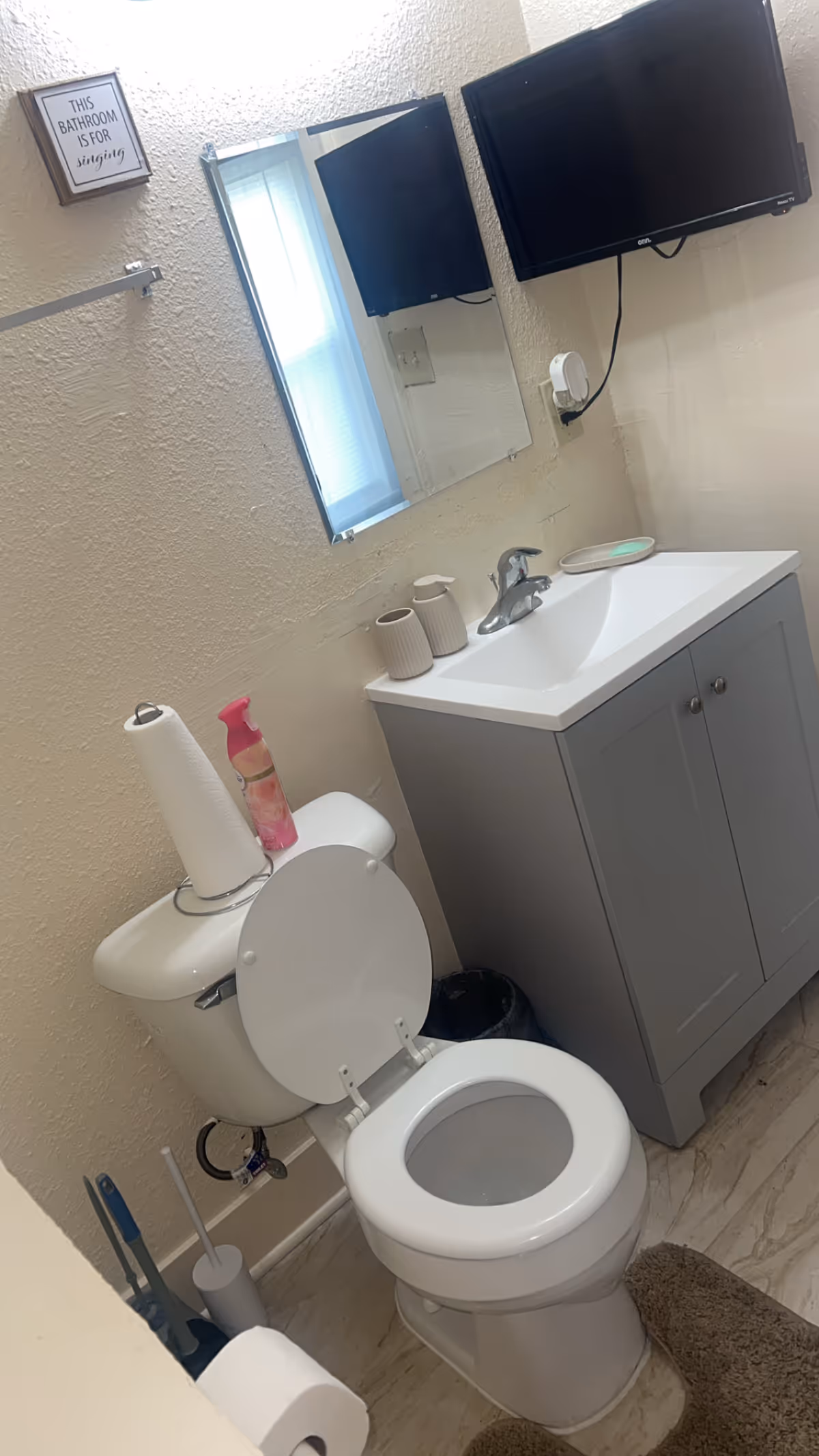 Small bathroom with toilet, sink vanity, wall-mounted TVs, and a sign on the textured wall.