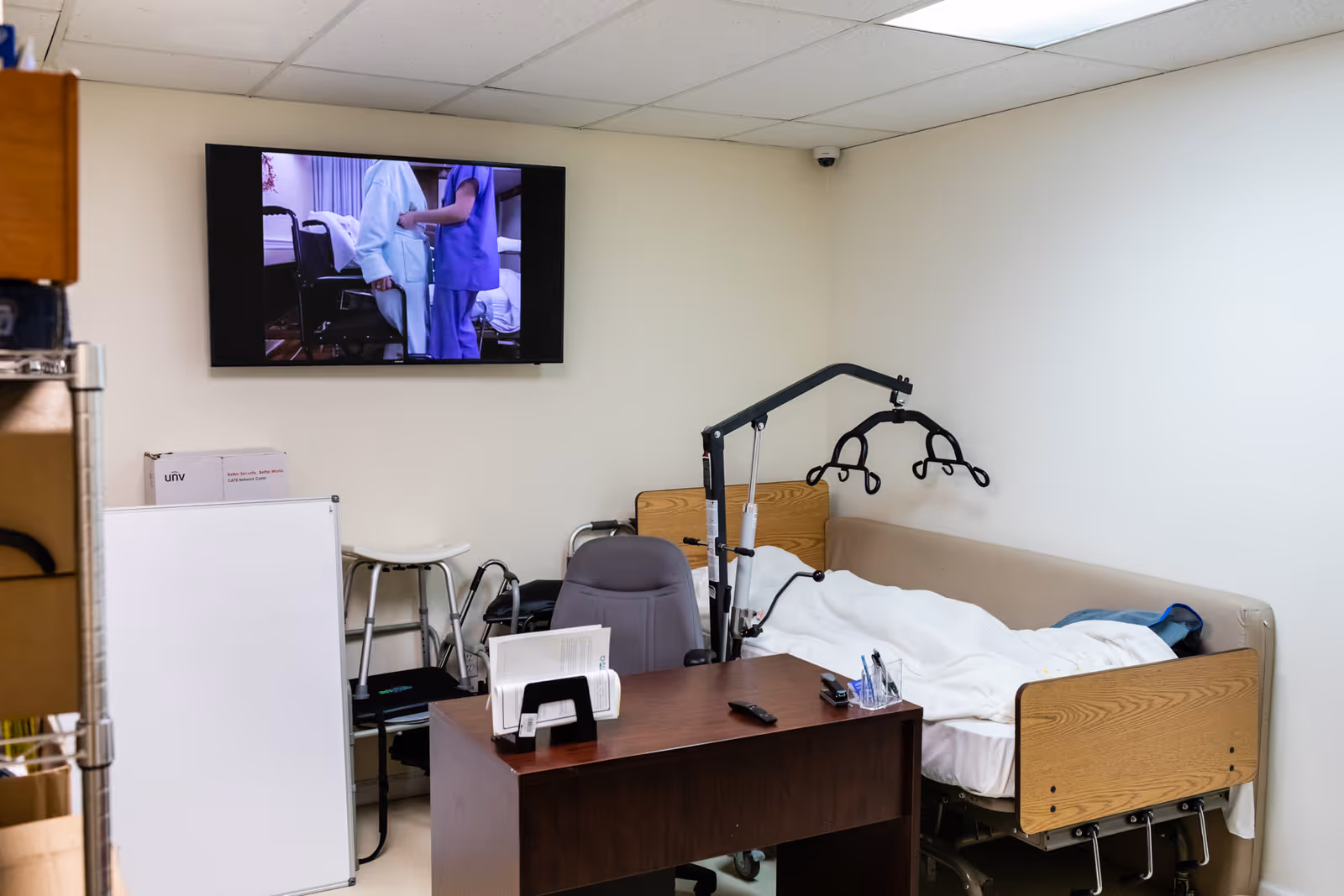 Hospital-style patient room with a bed, desk, chair, and wall-mounted monitor.