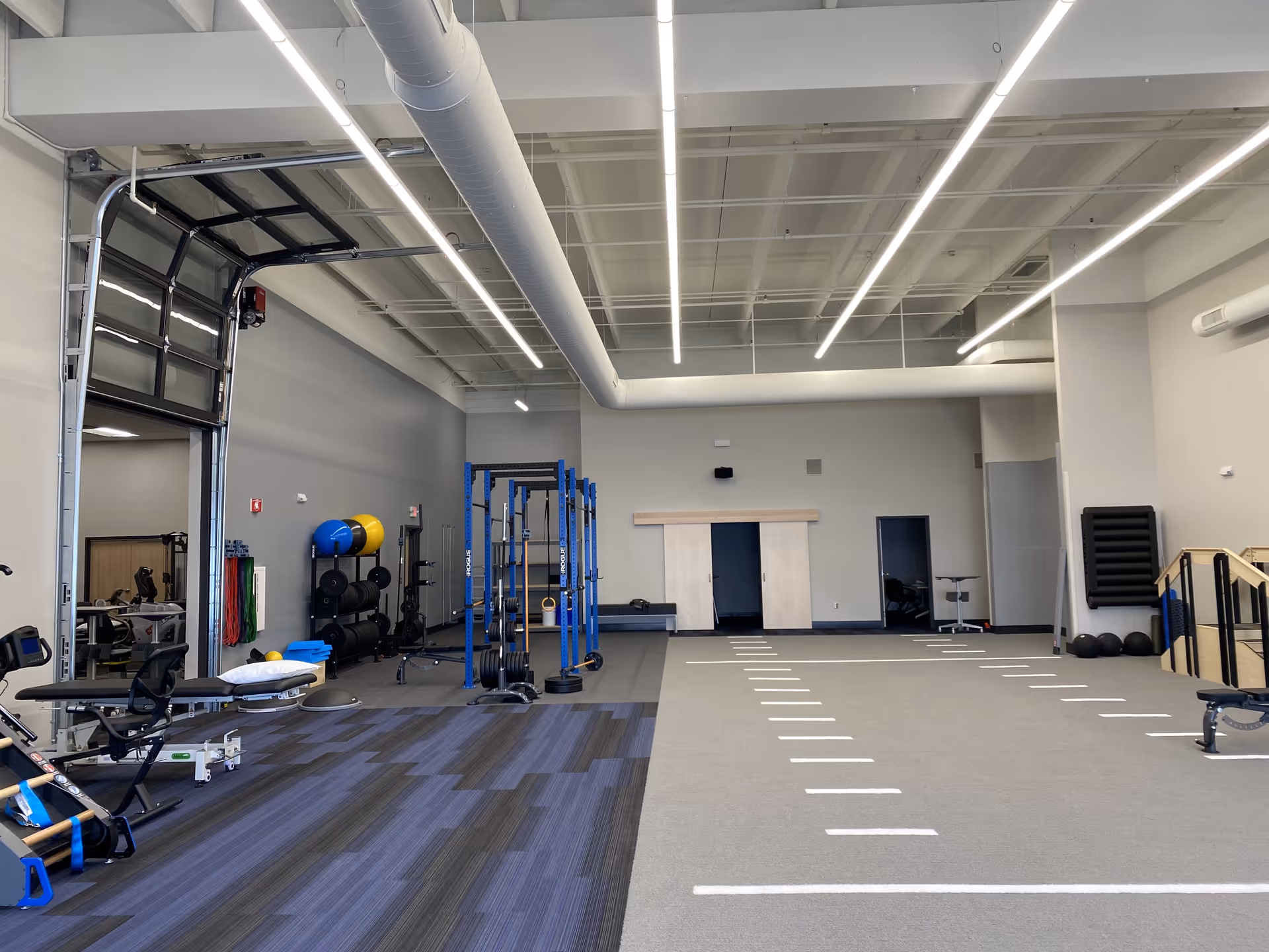 Spacious senior gym featuring blue weight racks, mats, and bright overhead lighting.