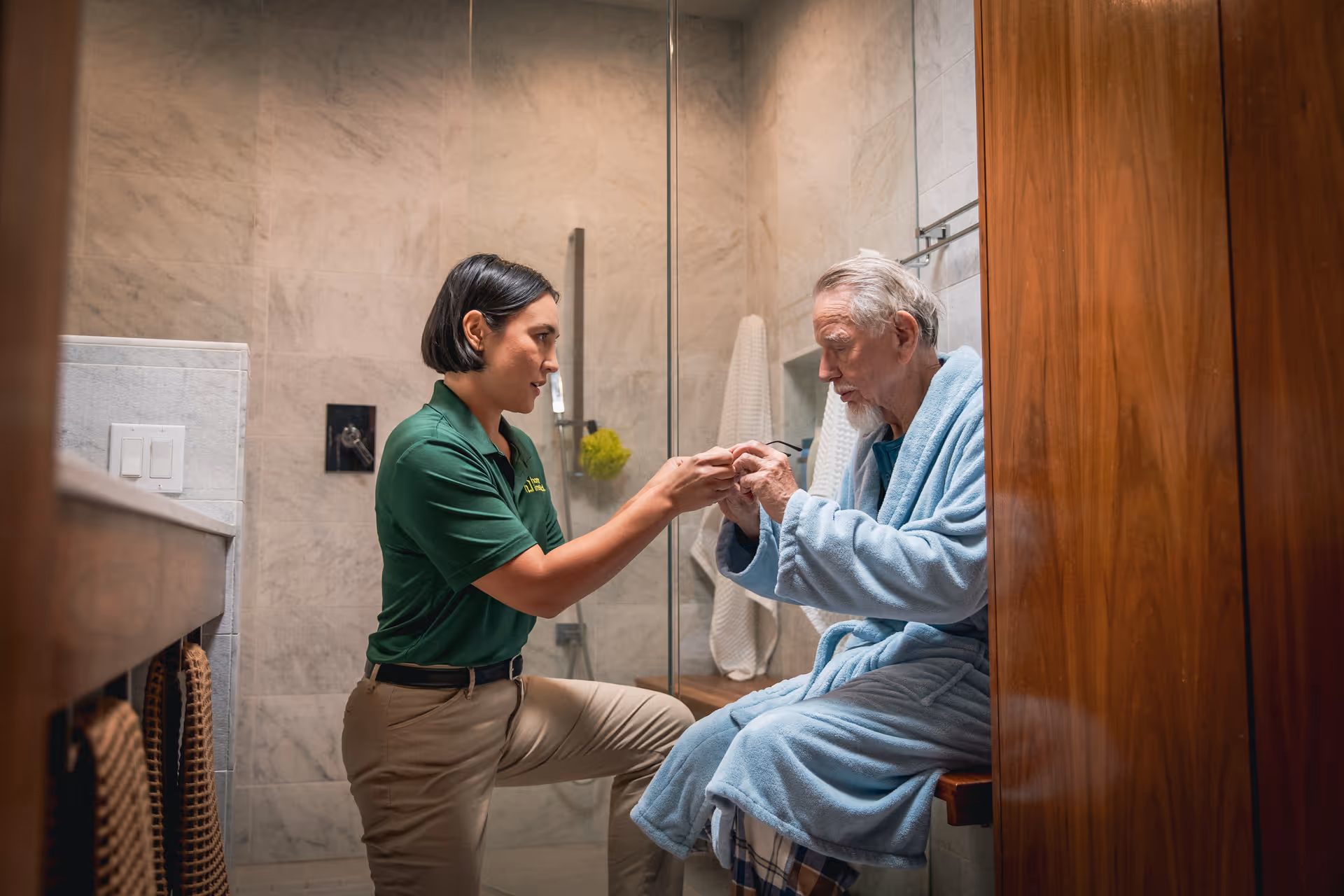 A caregiver assists an elderly man in a bathroom, holding his hands near the shower.
