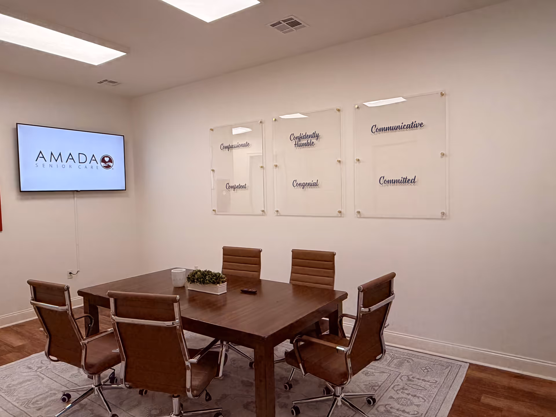 A modern conference room with a wooden table, brown chairs, and an Amada Senior Care screen.