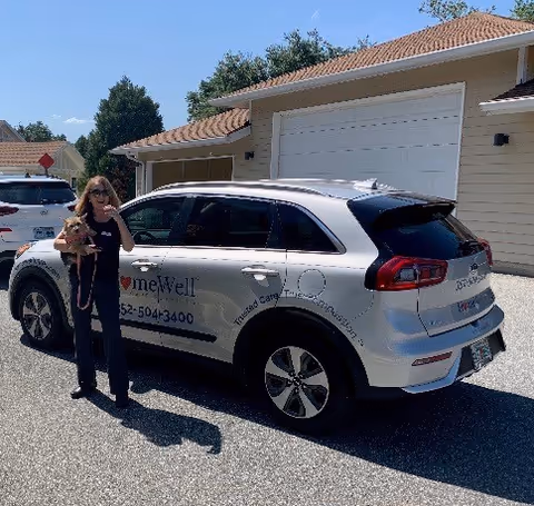 A woman stands beside a HomeWell Care Services SUV in a driveway with a small dog.