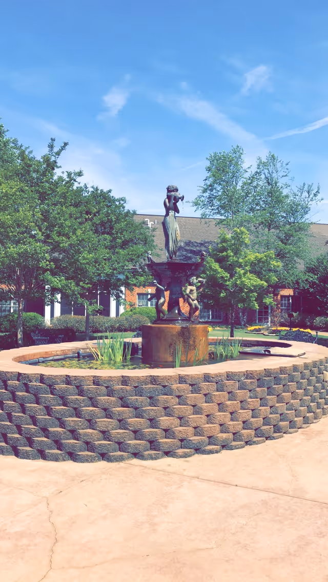 Outdoor courtyard with a tiered fountain statue surrounded by brick planter and trees.