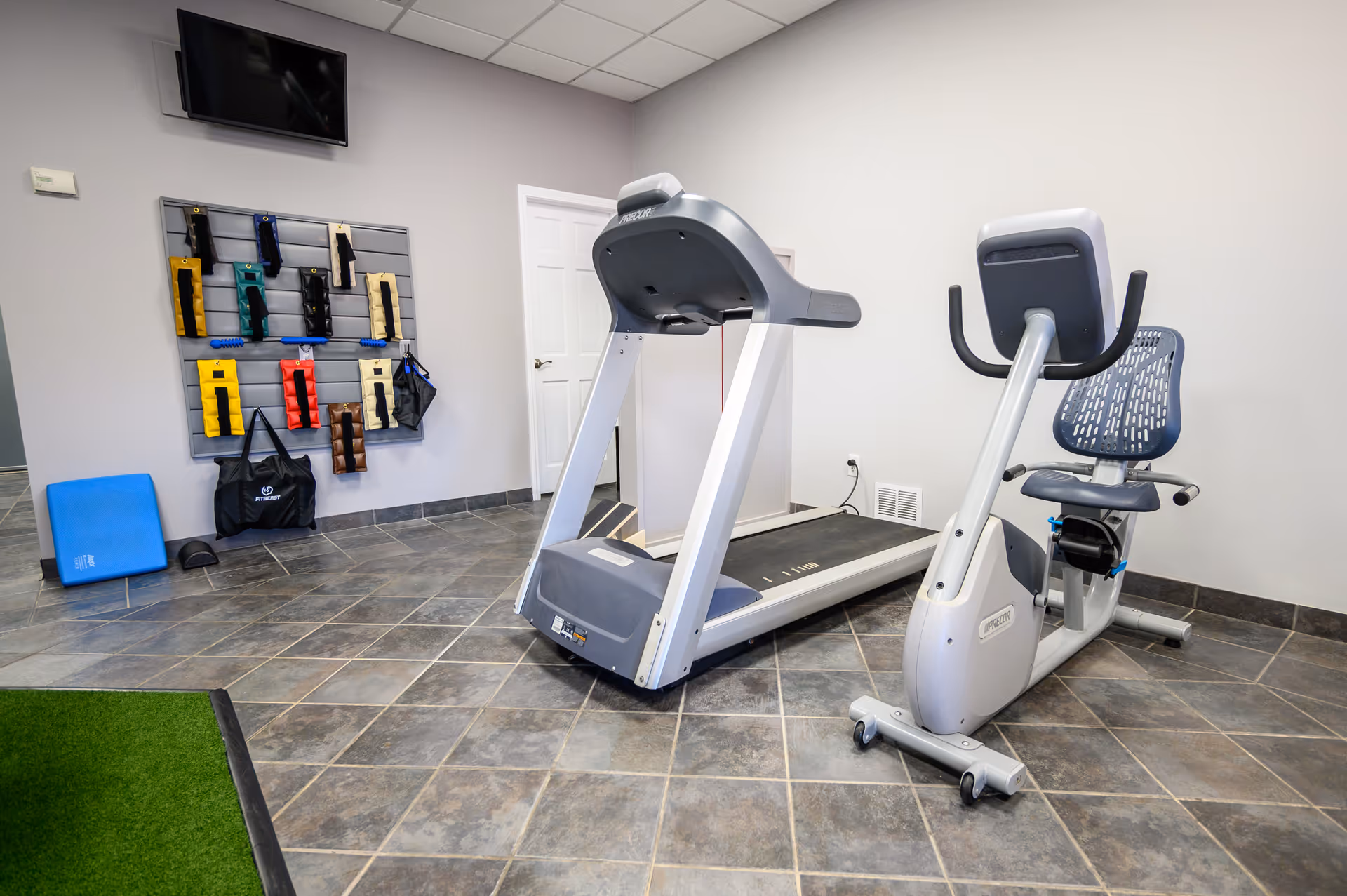 Small fitness room with treadmill, stationary bike, wall accessories, and tiled floor.