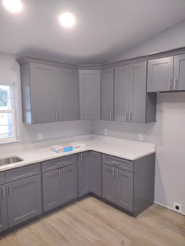 Gray cabinets surround an L-shaped kitchen with white countertops and a sink.