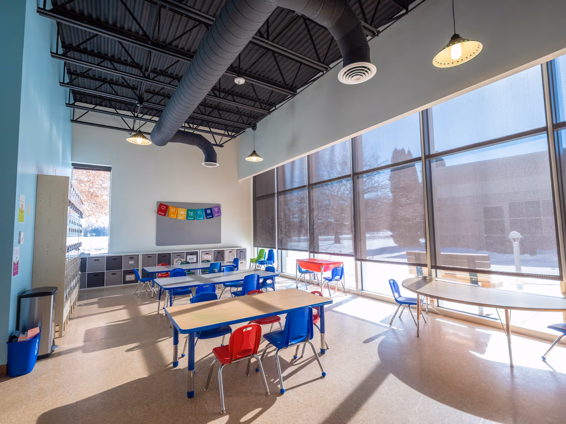 Bright classroom-like activity room with blue chairs, tables, lockers, and large windows.