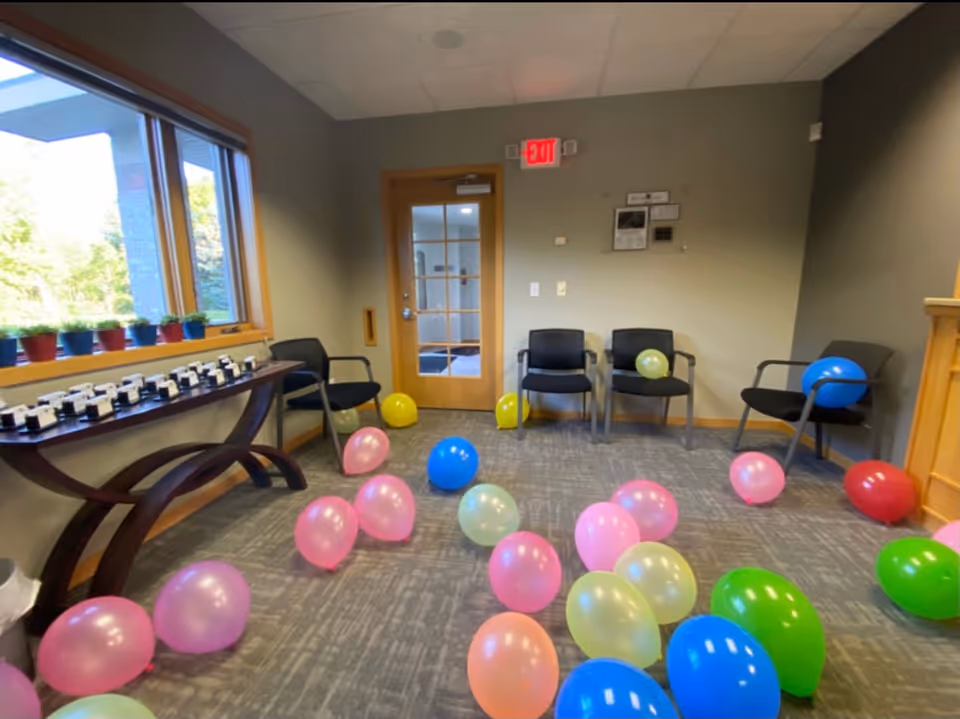 A waiting or lounge room with colorful balloons scattered on the carpet and chairs along walls.