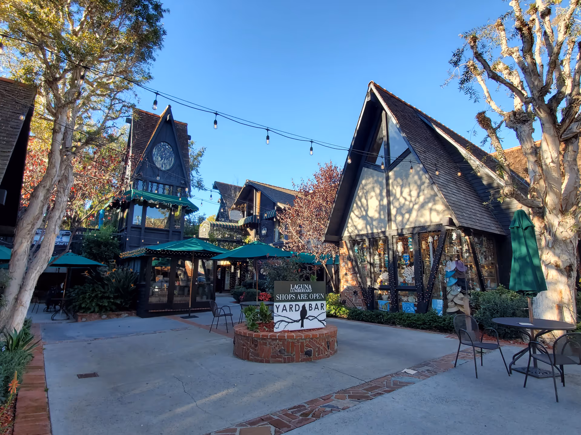 Outdoor courtyard with A-frame storefronts, string lights, and green umbrellas.
