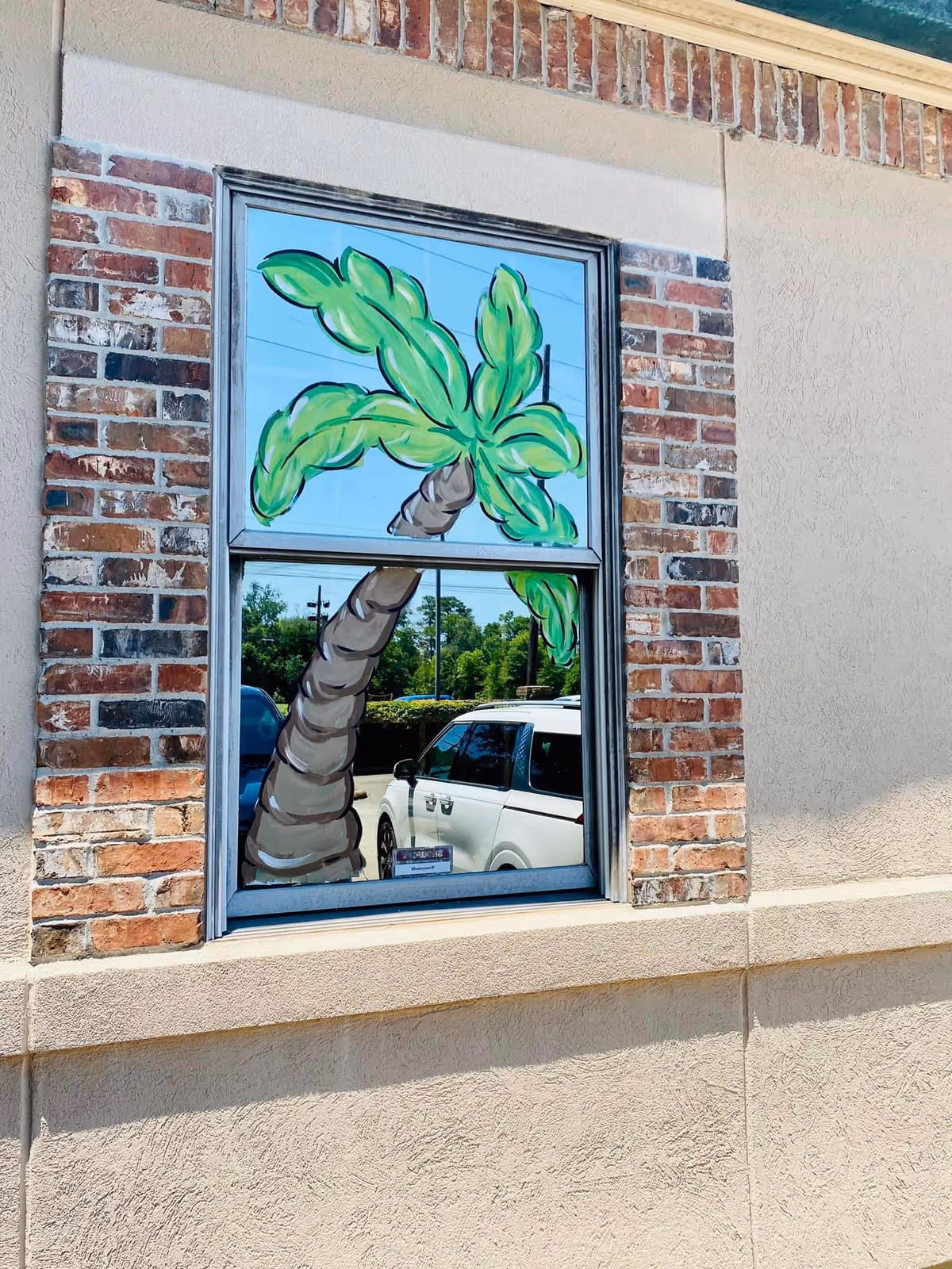 Exterior brick wall window features a painted tropical palm mural with a reflected parked car.