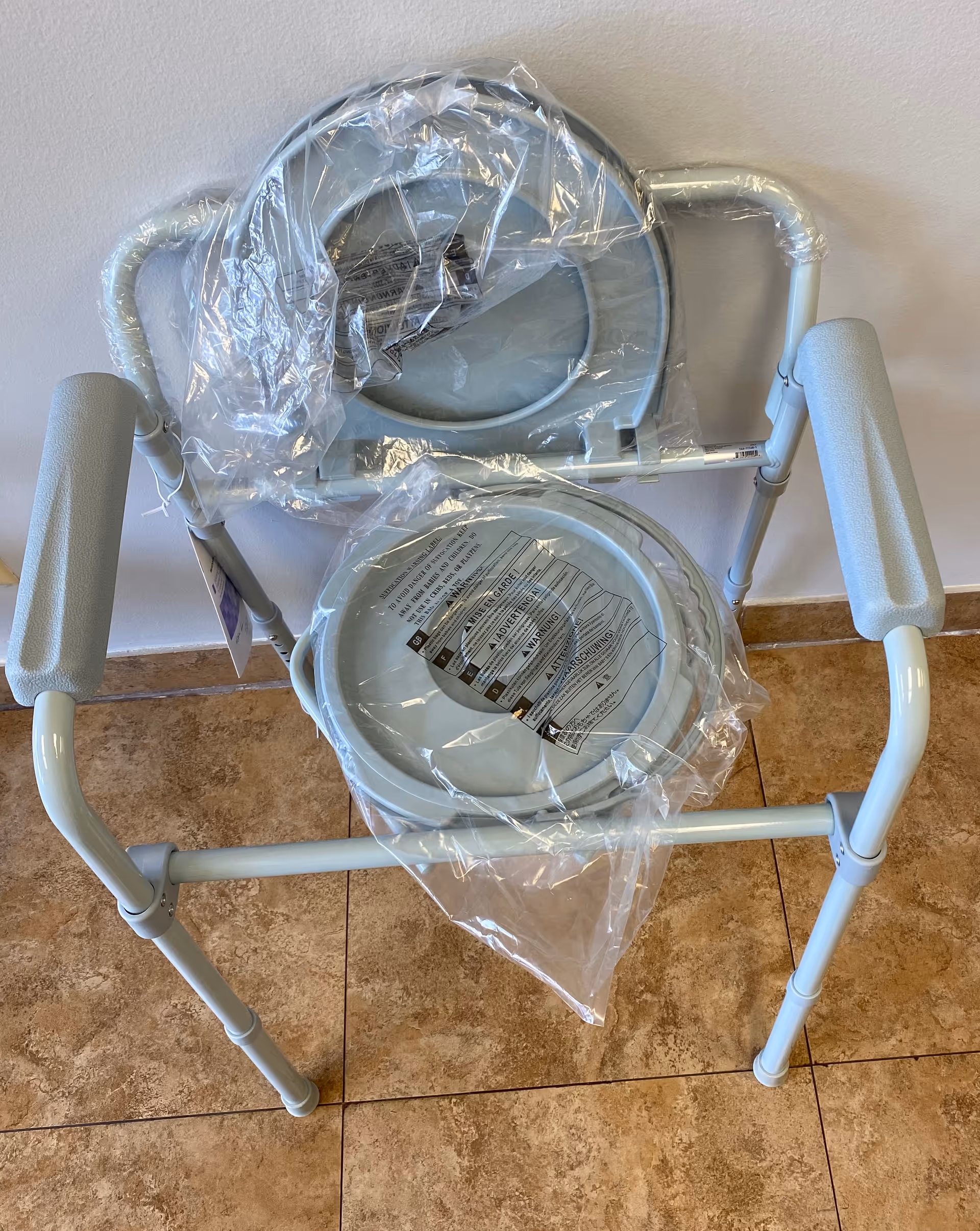 Plastic-wrapped medical commode chair sits in a tiled interior.