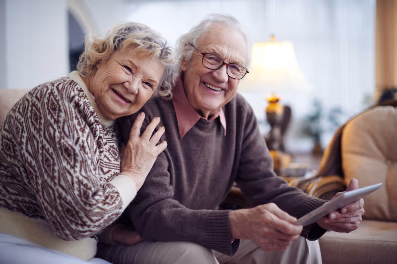 Two elderly people sit together on a sofa in a living room, smiling.