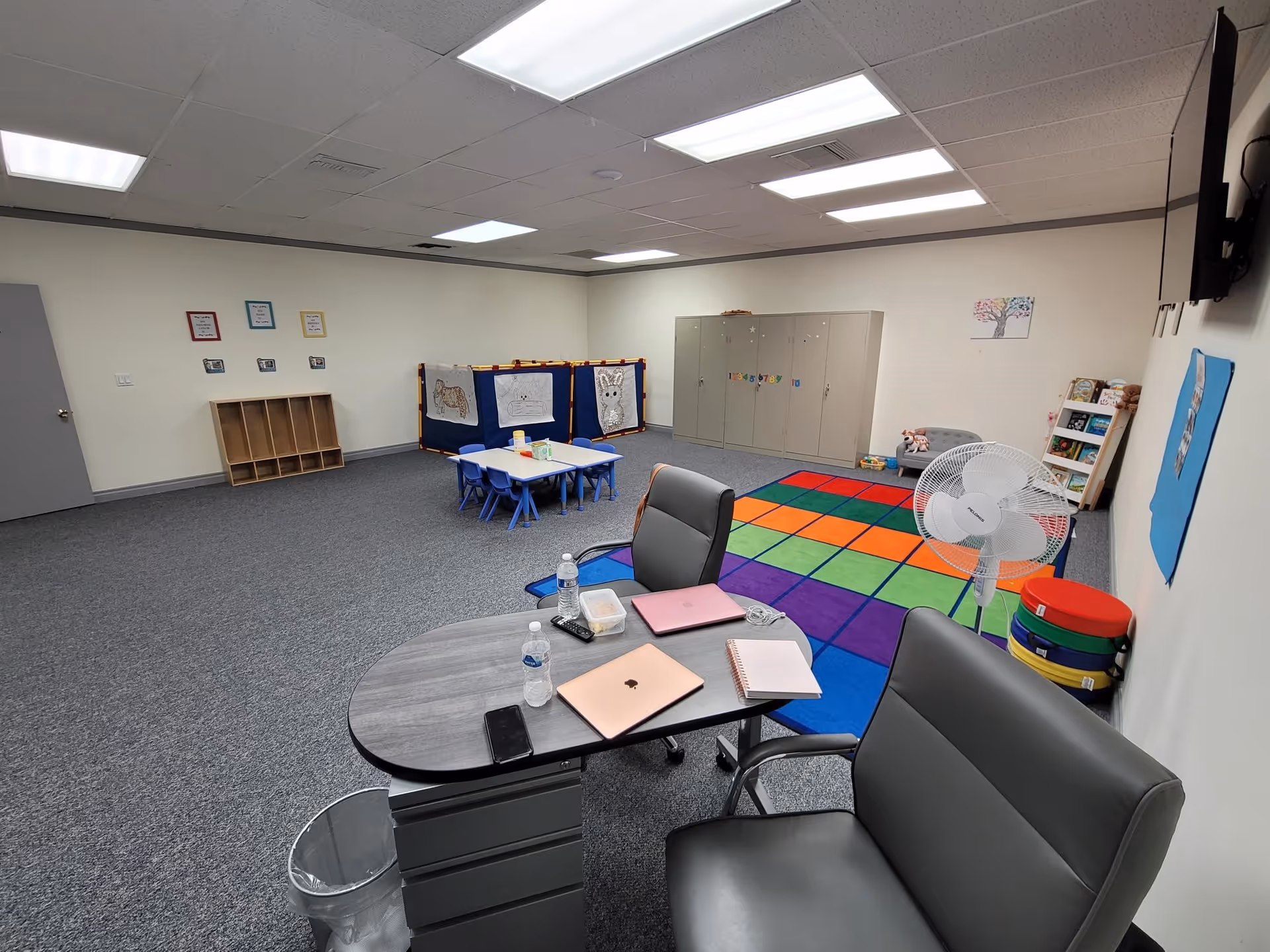Interior activity room with colorful rug, small blue chairs, lockers, and a desk.