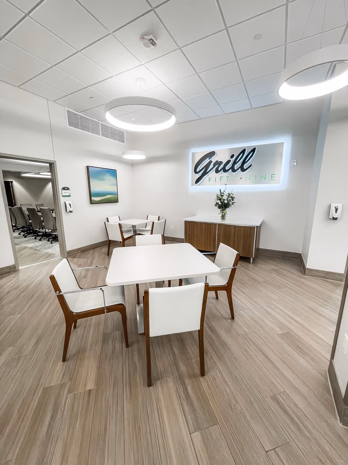 Modern dining area with white tables, wooden chairs, and a lit 'Grill Fifty-Nine' sign.