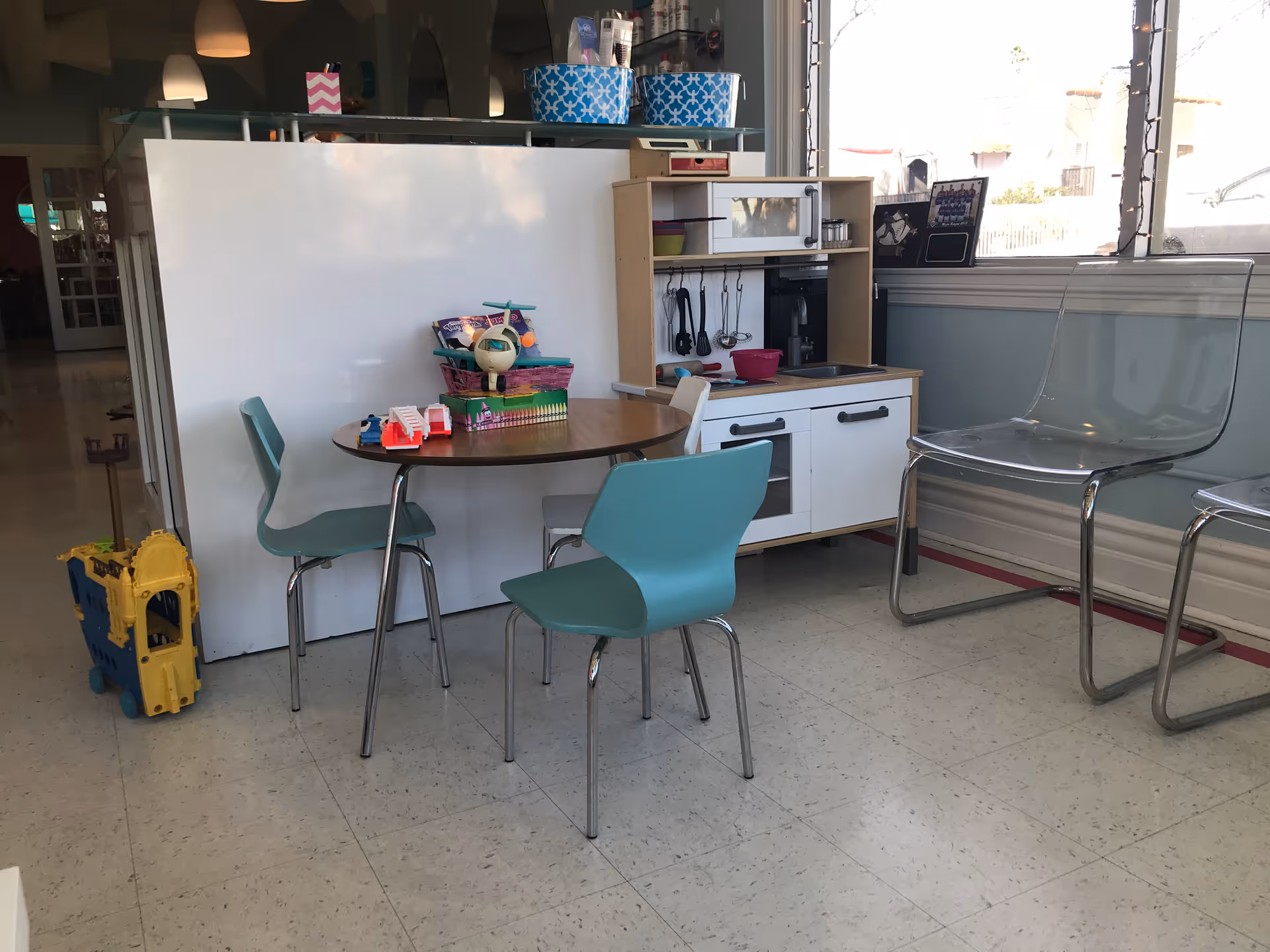 Children's play kitchen area with small table, turquoise chairs in a bright room.