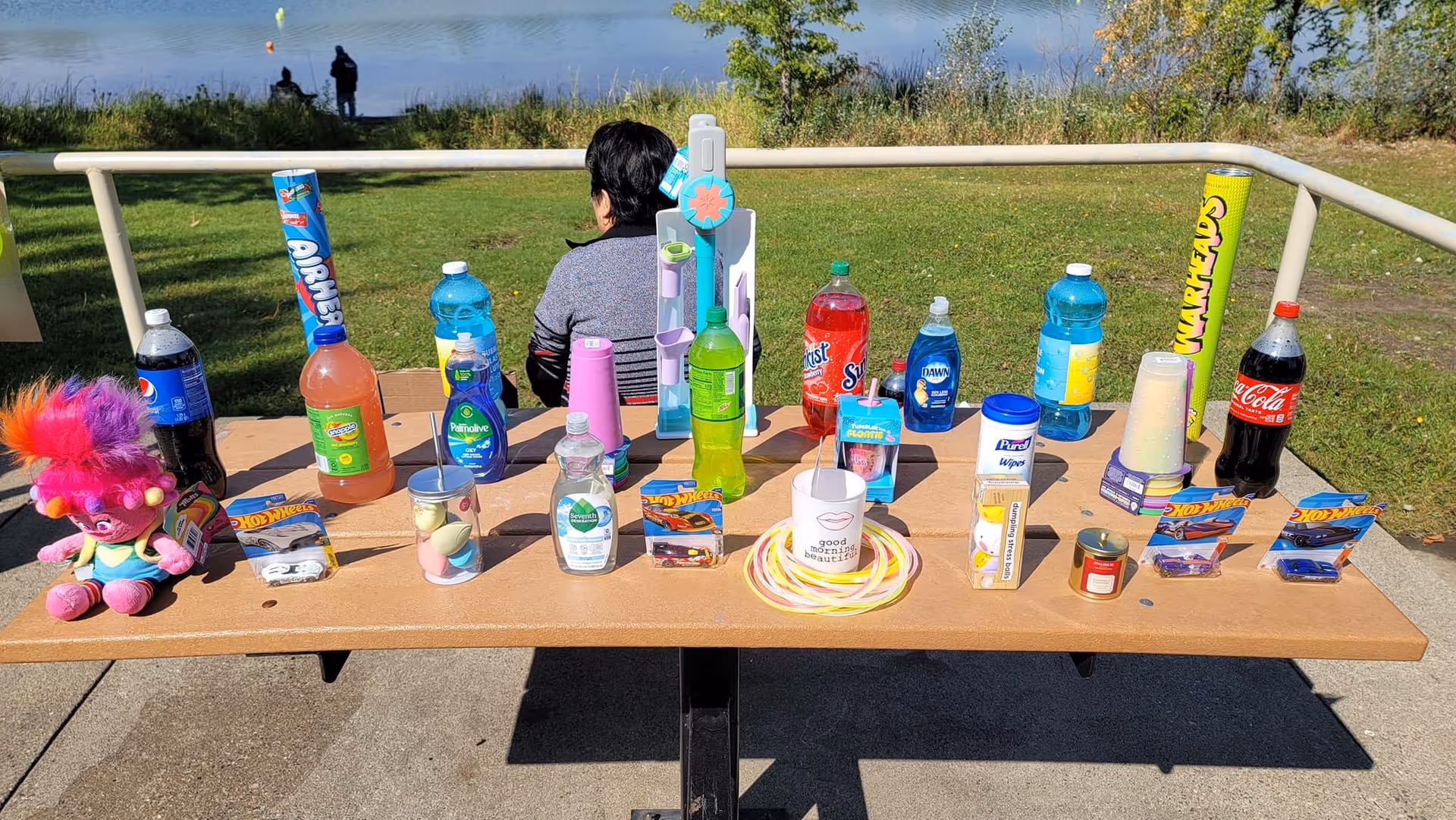 Outdoor table displays colorful beverages, toys, and miscellaneous items; a person stands in the background.