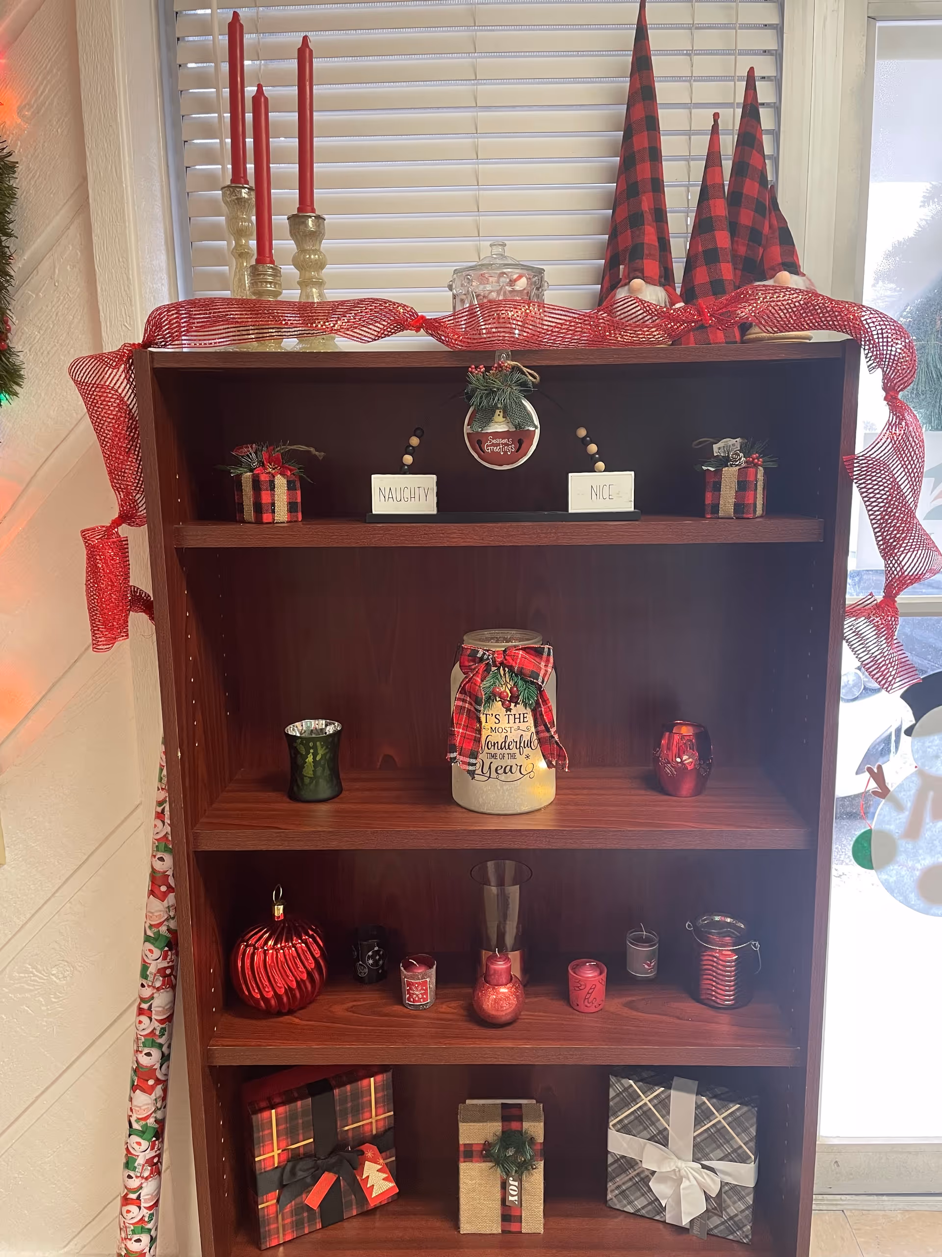 Wood bookshelf decorated with red holiday decor and signs beside a window with blinds.