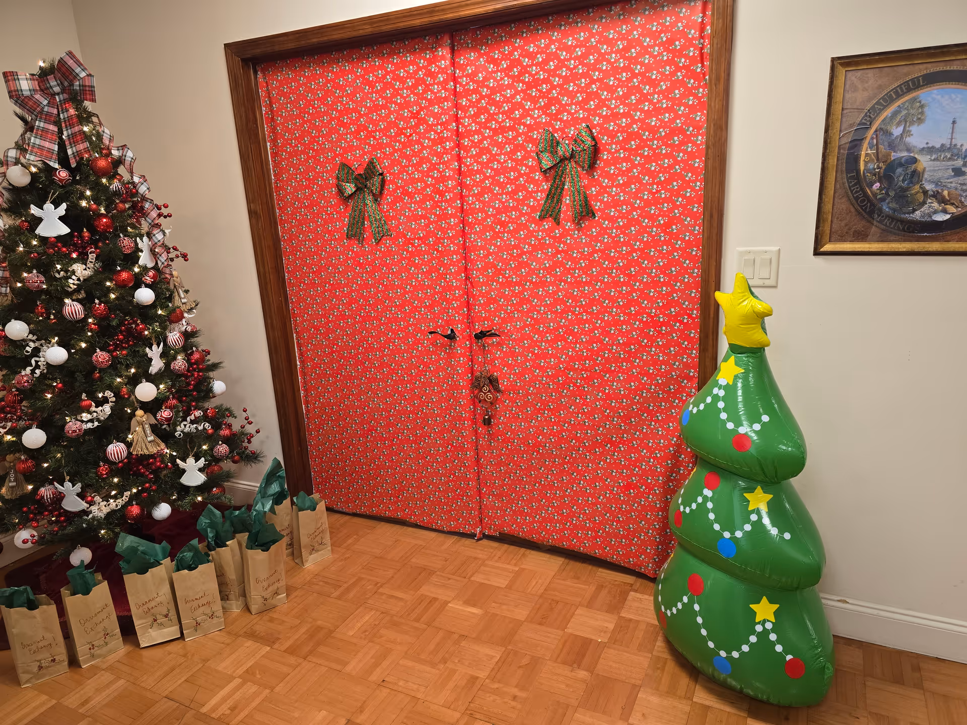 A Christmas tree and gift bags sit beside red patterned double doors in a decorated hallway
