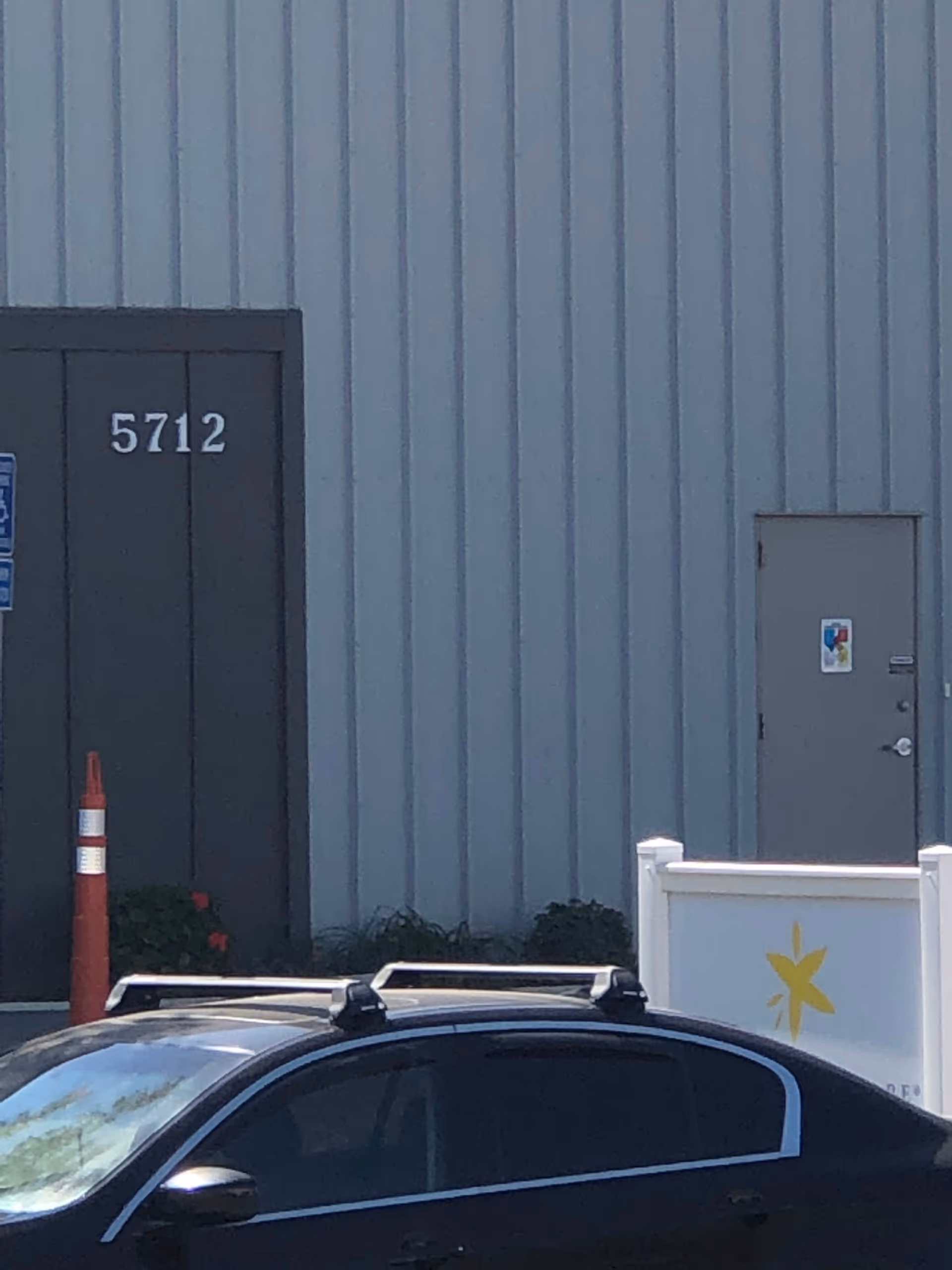 Exterior view of a gray metal building with numbered door, cone, fence, and parked car.