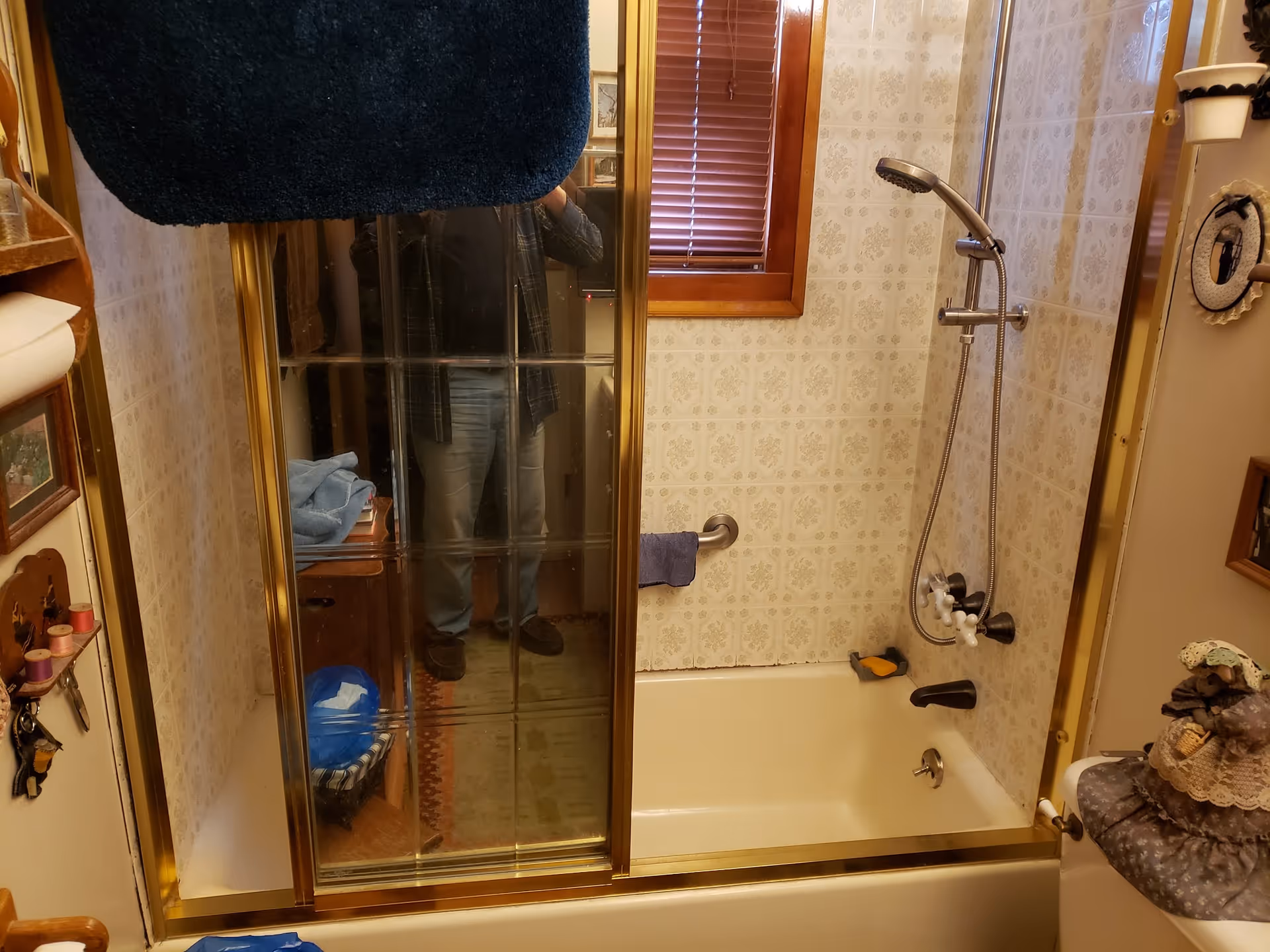 Shower-tub combo with glass doors, brass frame, patterned walls, and a reflection of a person.
