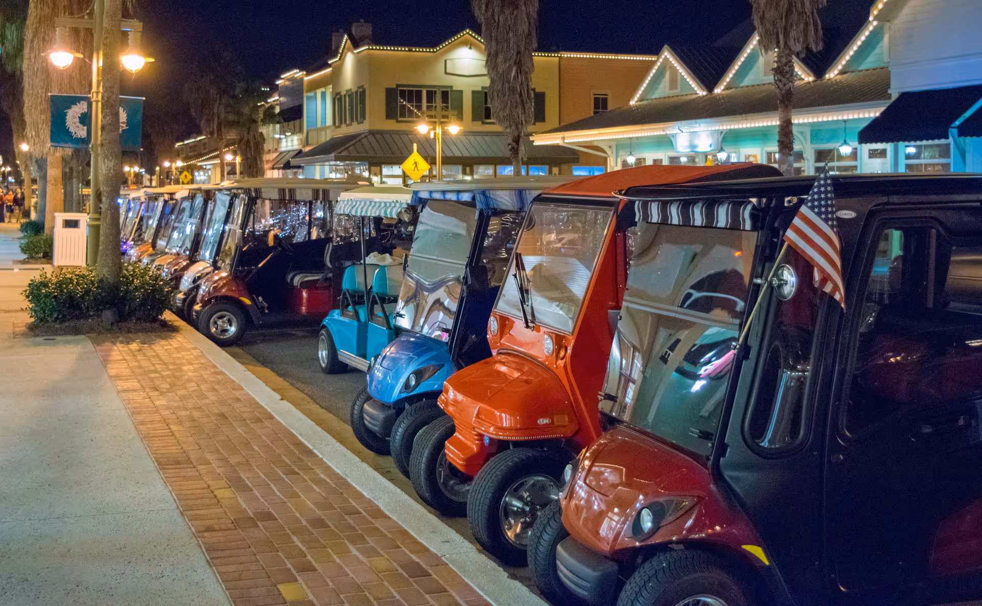 Row of colorful golf carts lined up along a sidewalk at night near storefronts.