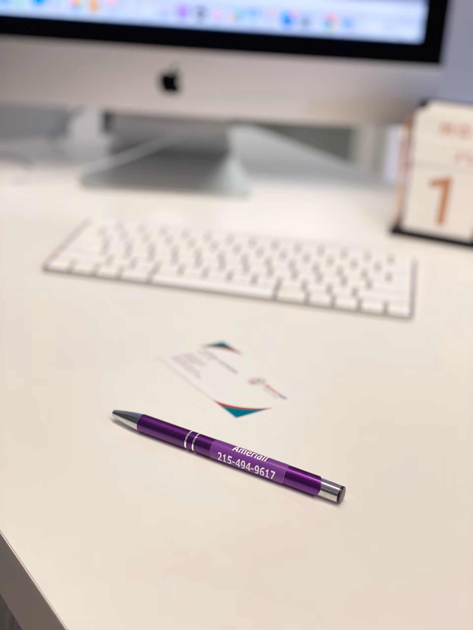 An office desk with a white keyboard, iMac monitor, and purple promotional pen.