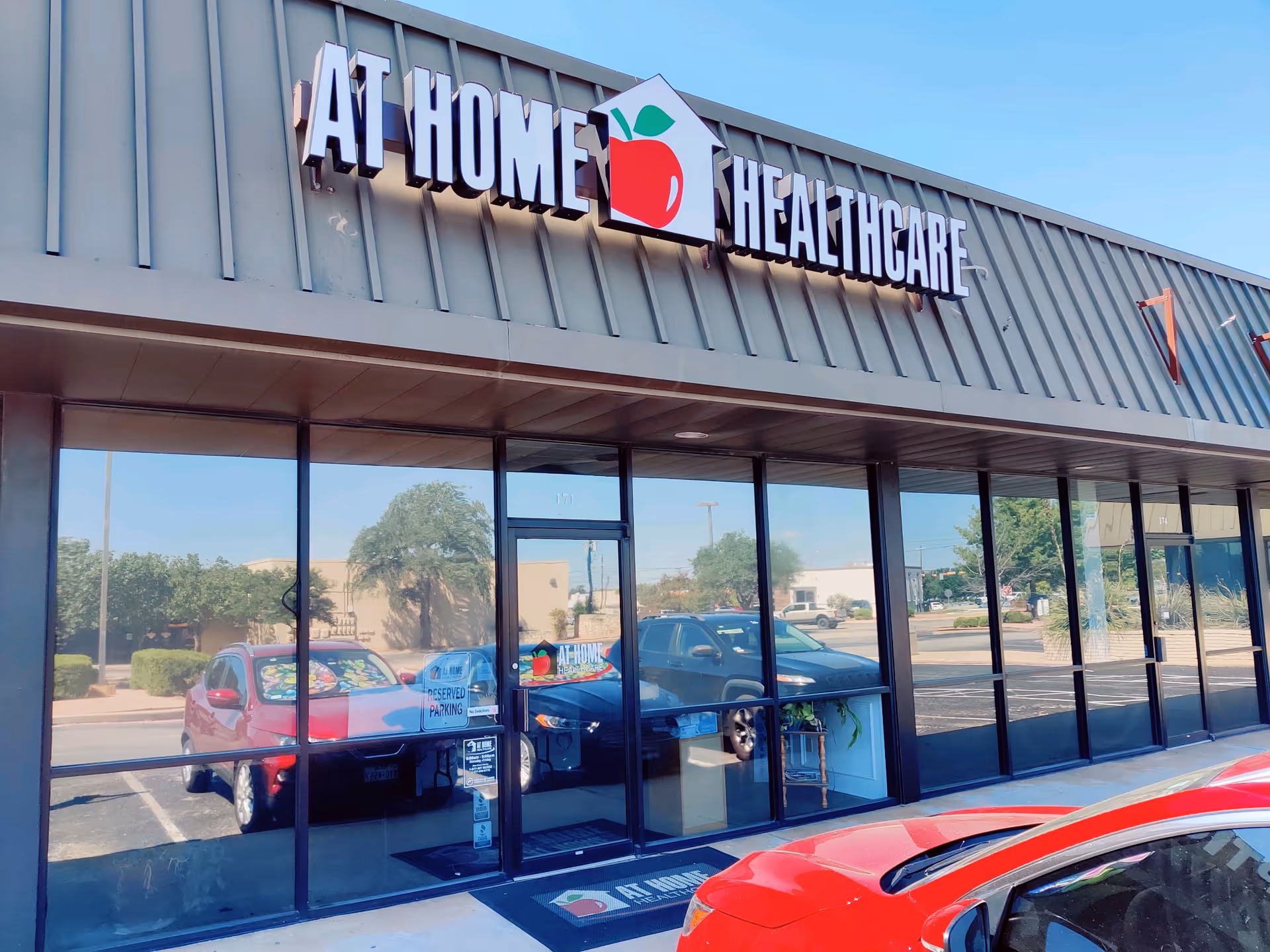 Exterior view of At Home Healthcare storefront with glass doors and parked cars.