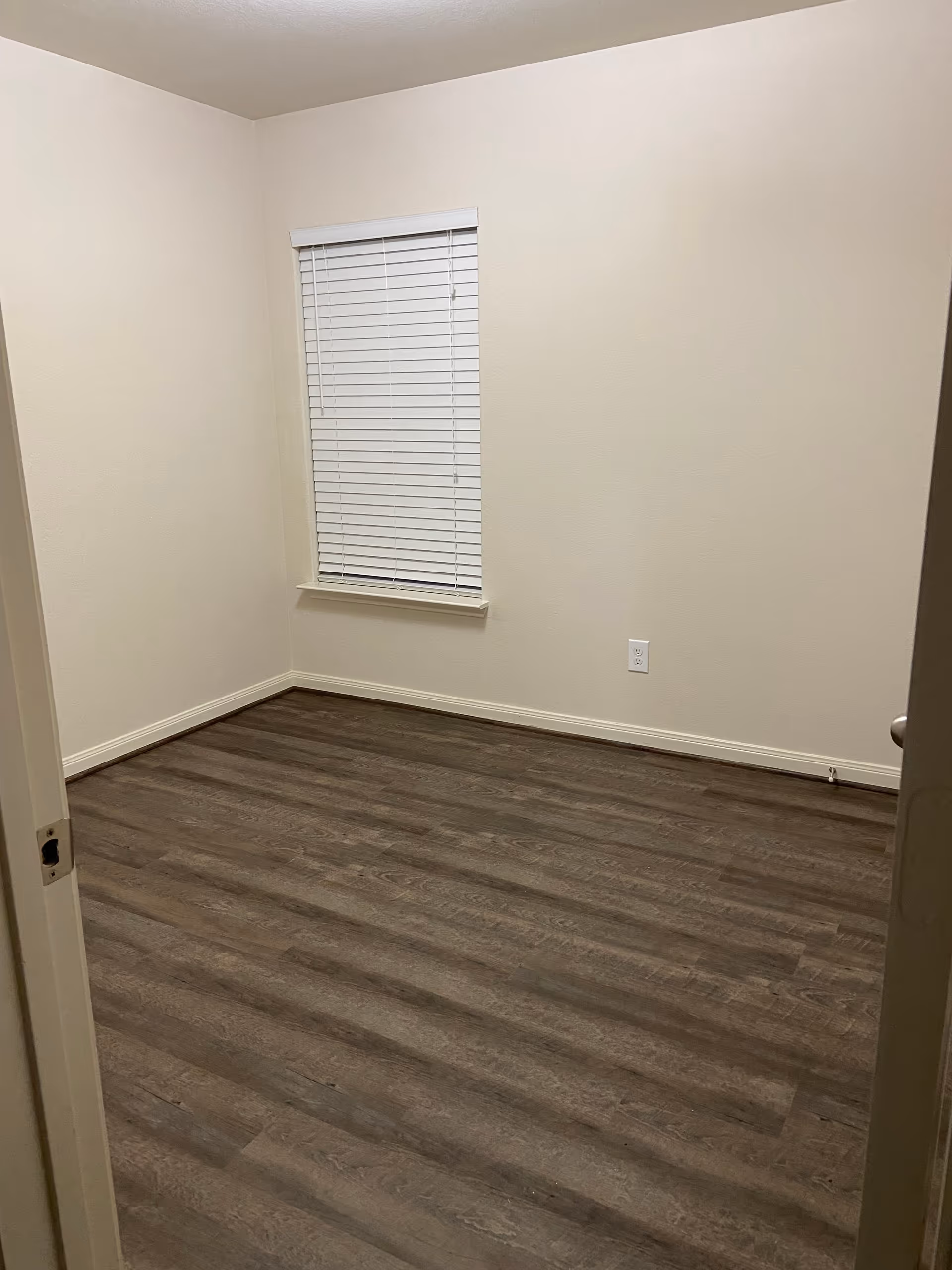 Empty beige room with a window and blinds, wood-like flooring, in a residential interior.