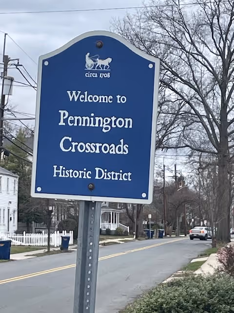 Blue welcome sign for Pennington Crossroads Historic District beside a residential street.