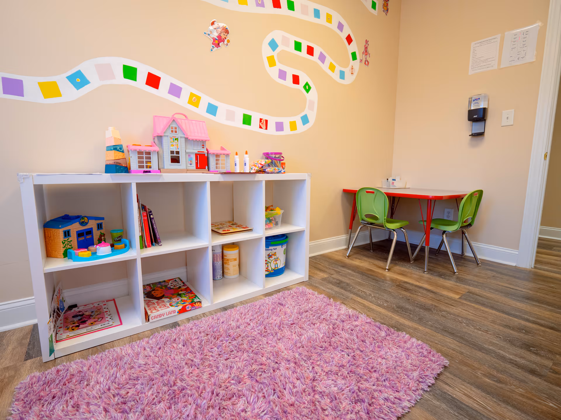 Play corner with toy shelves, pink shag rug, and kid-sized table with green chairs.