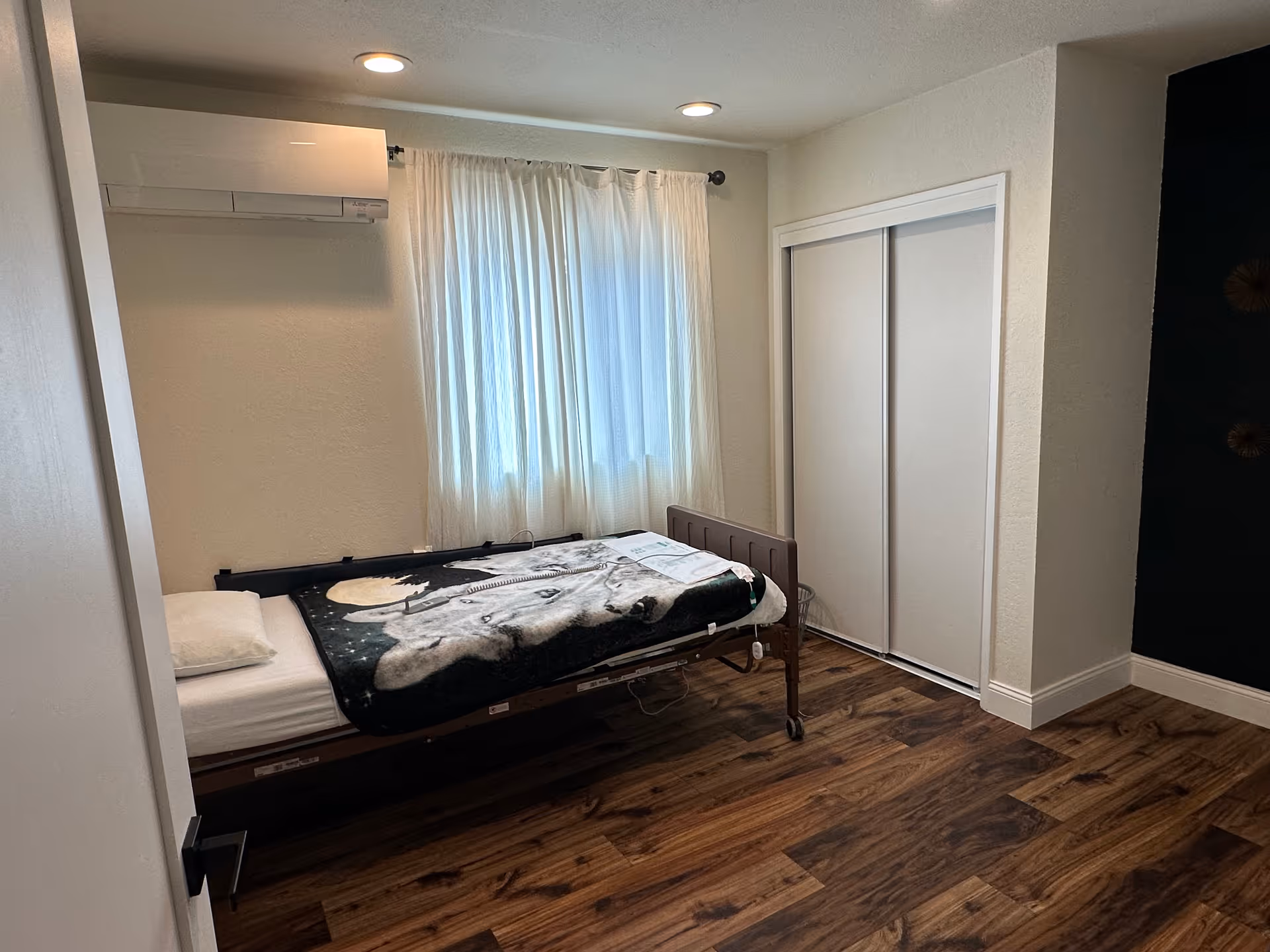 A single bedroom in a senior facility with an adjustable bed, window curtains, and closet.