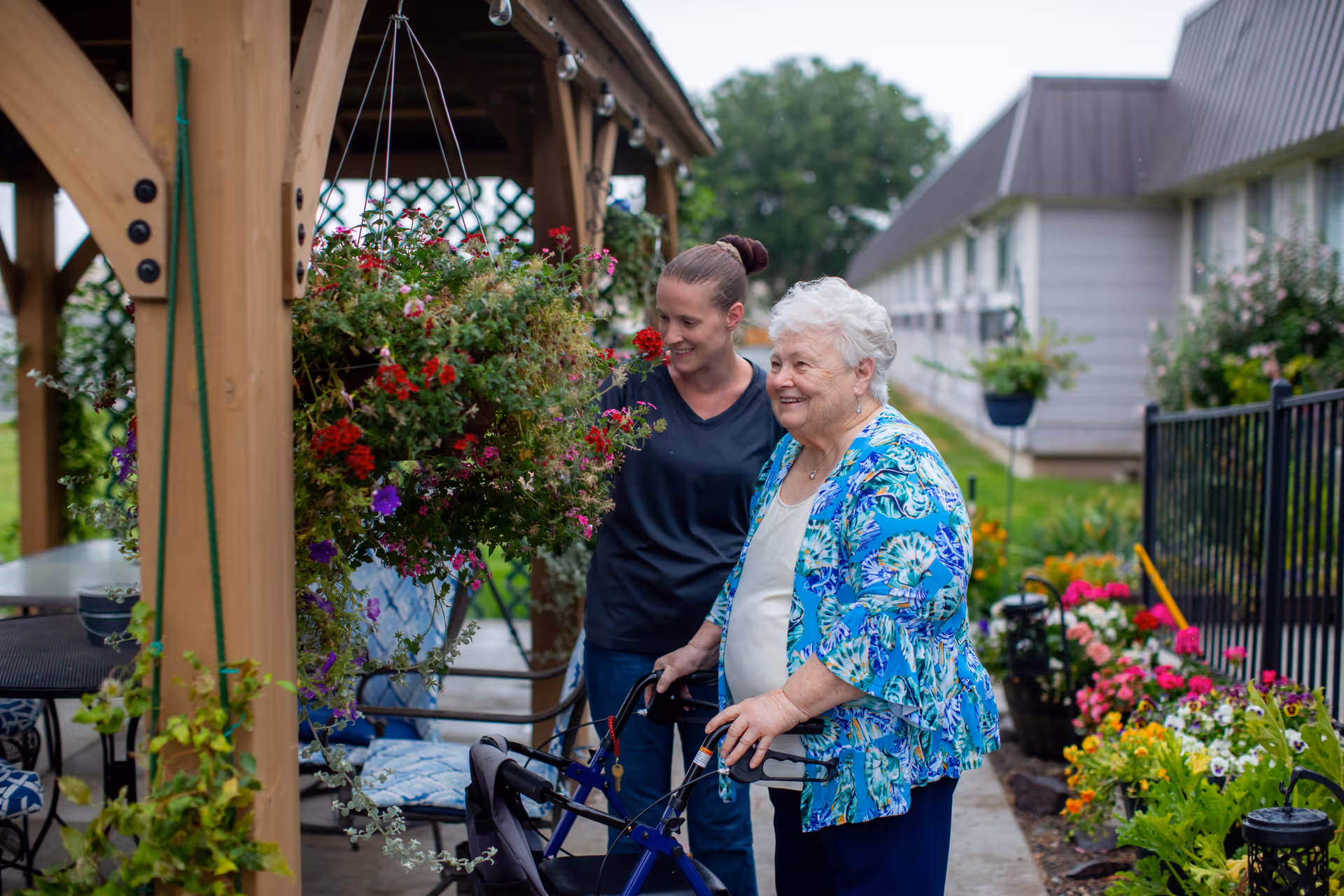 An elderly woman using a walker is guided by a caregiver in a flower-filled outdoor garden.