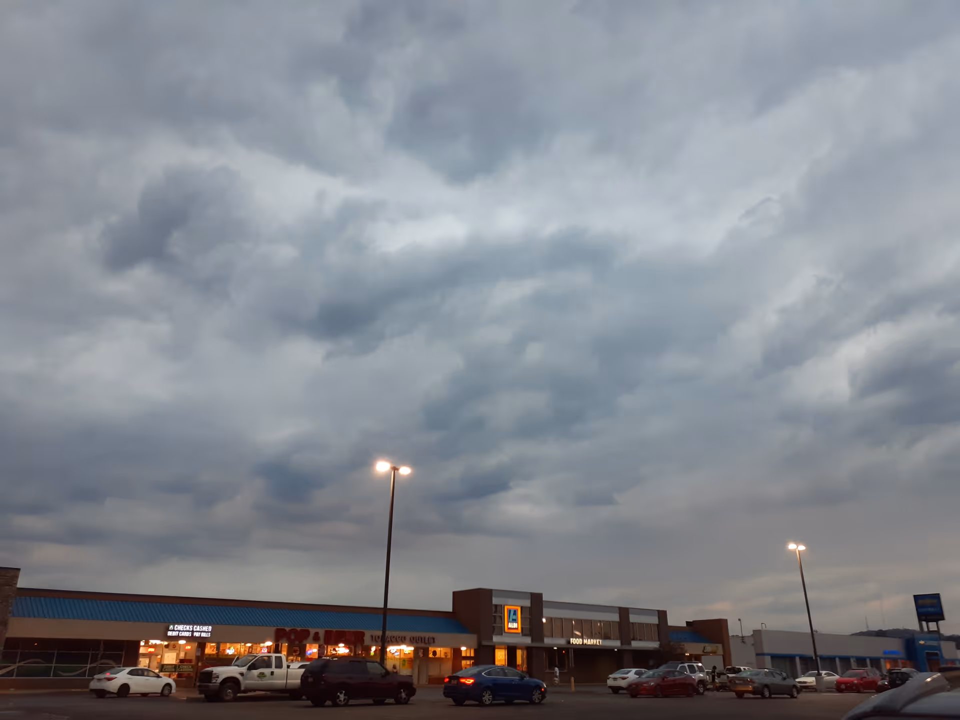 Overcast sky over a strip mall parking lot with storefronts and parked cars.