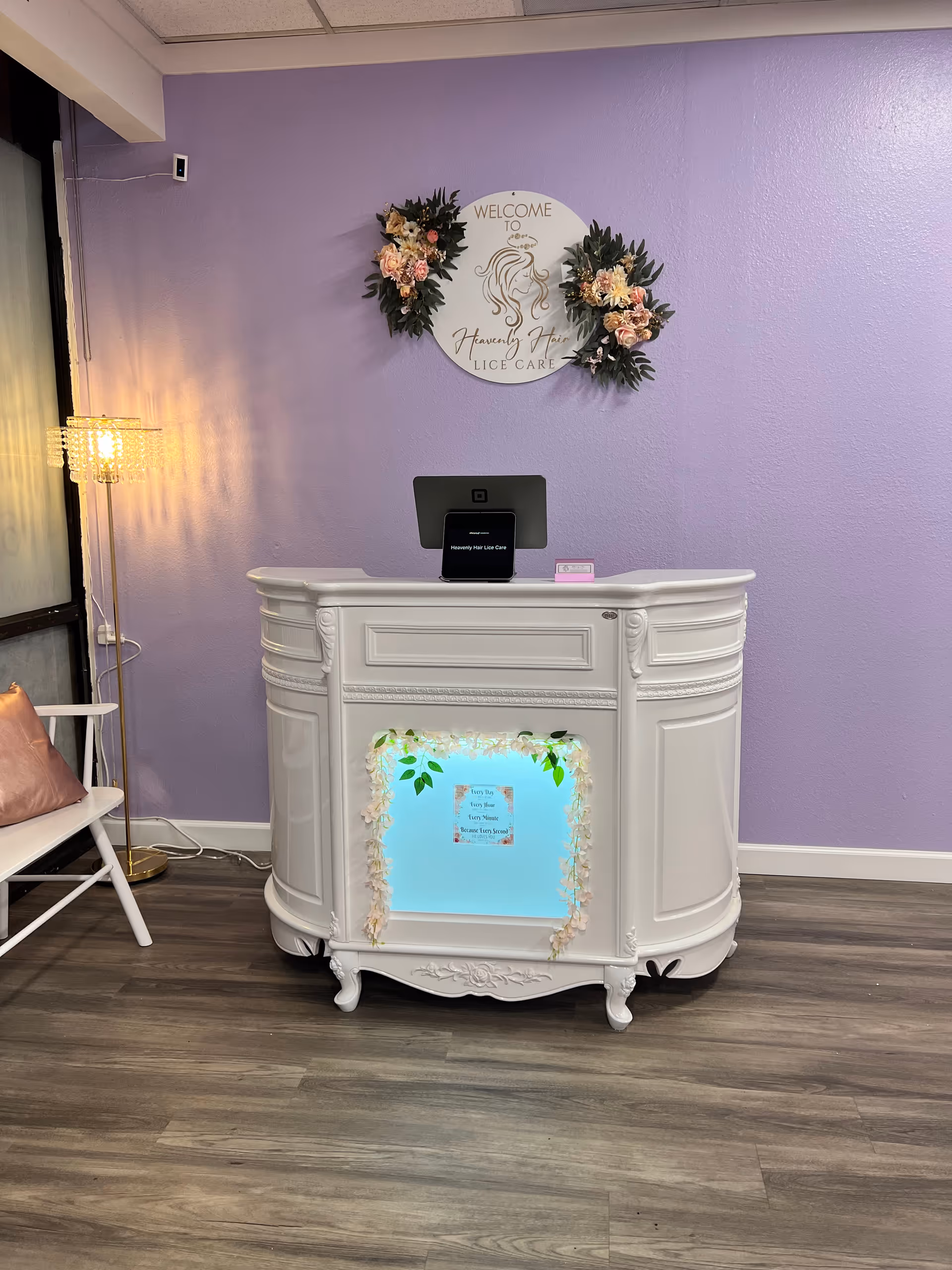 White ornate reception desk against lavender wall with sign reading 'Welcome to Heavenly Hair Lice Care'.