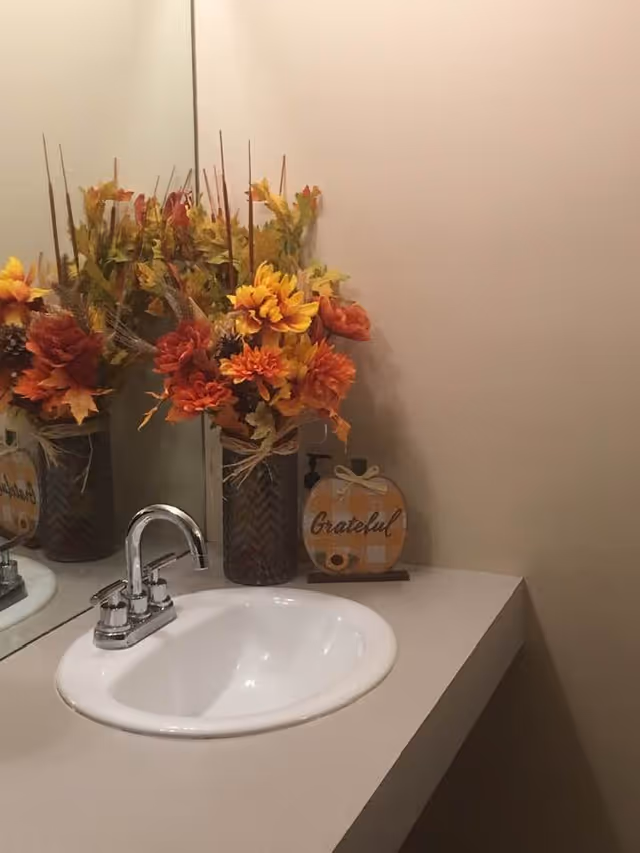 Bathroom vanity with sink, mirror, autumn flowers in vases, and a Grateful sign.