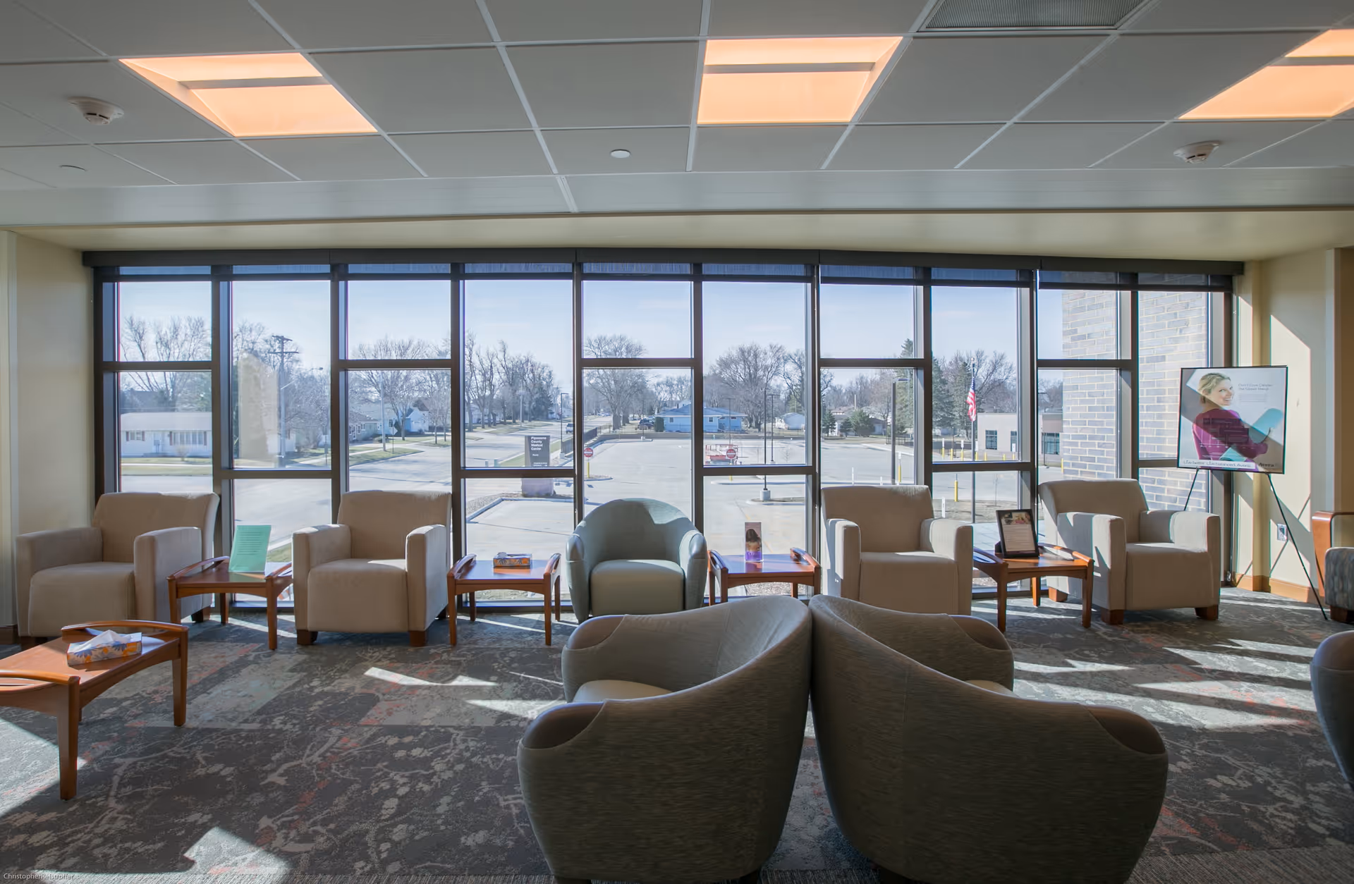 Bright hospital lobby lounge with multiple beige chairs, small wooden tables, and large windows.